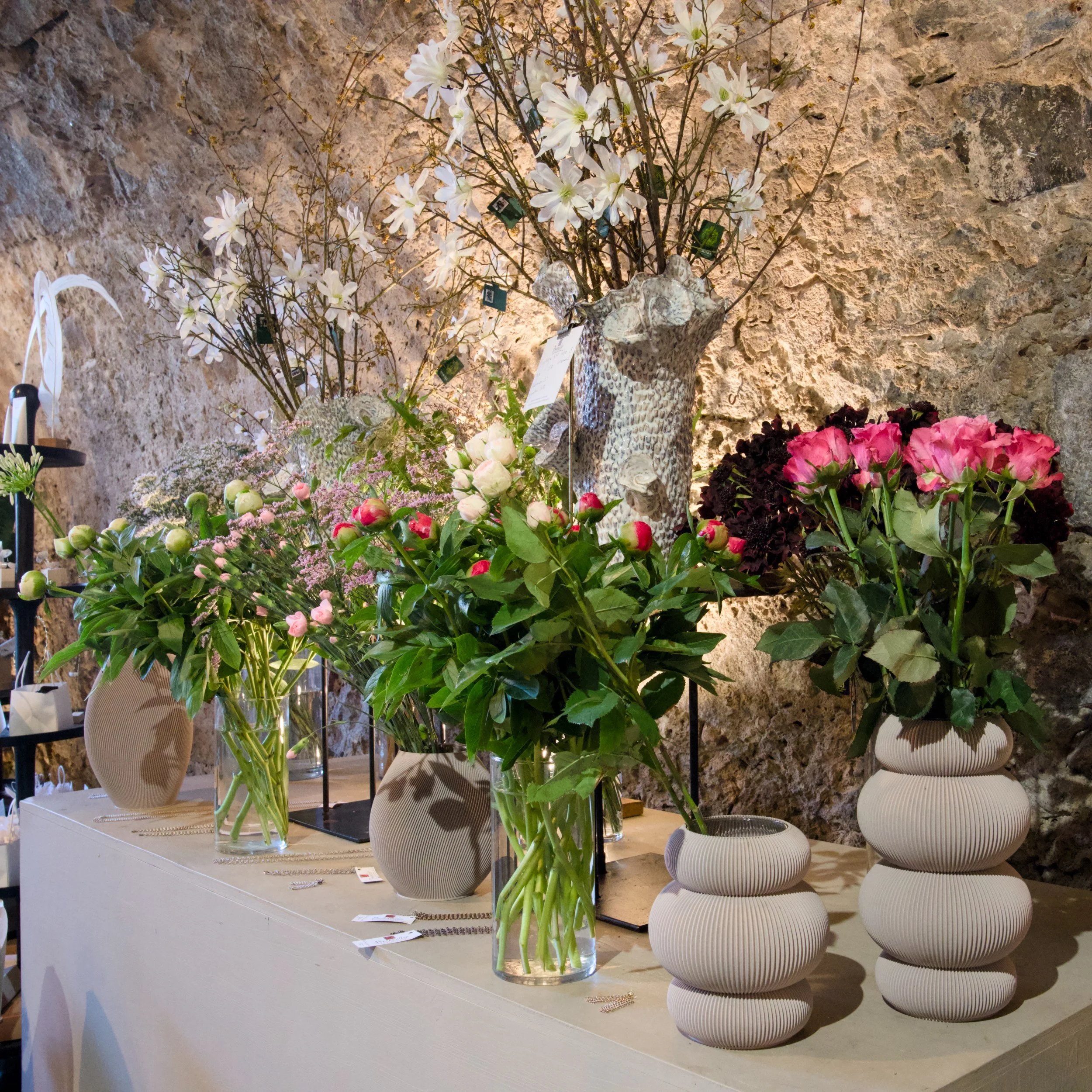 Blumen in Vasen auf weißem Tisch vor einer Steinwand.