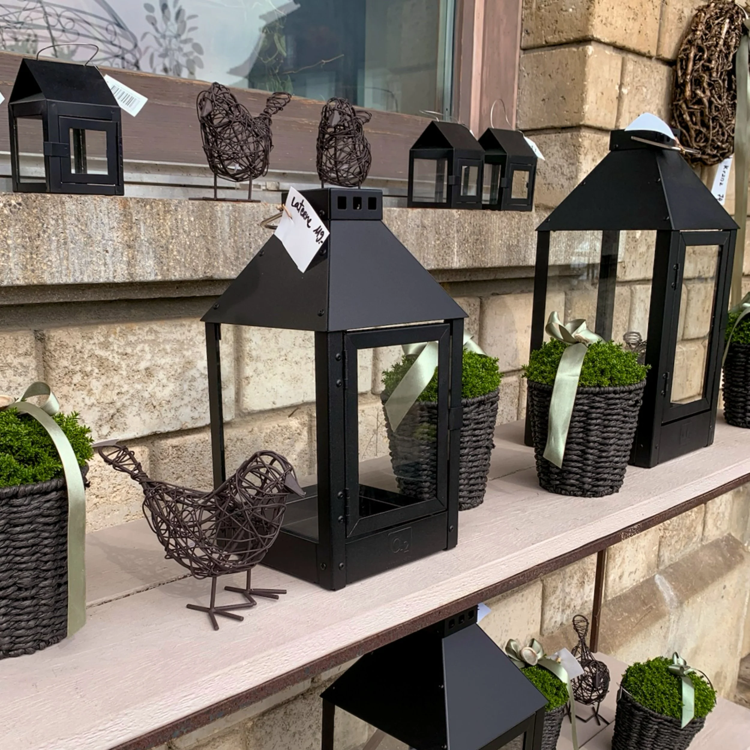 Dekorative Gartenlampen und Pflanzen auf einem Regal vor einer Steinmauer.