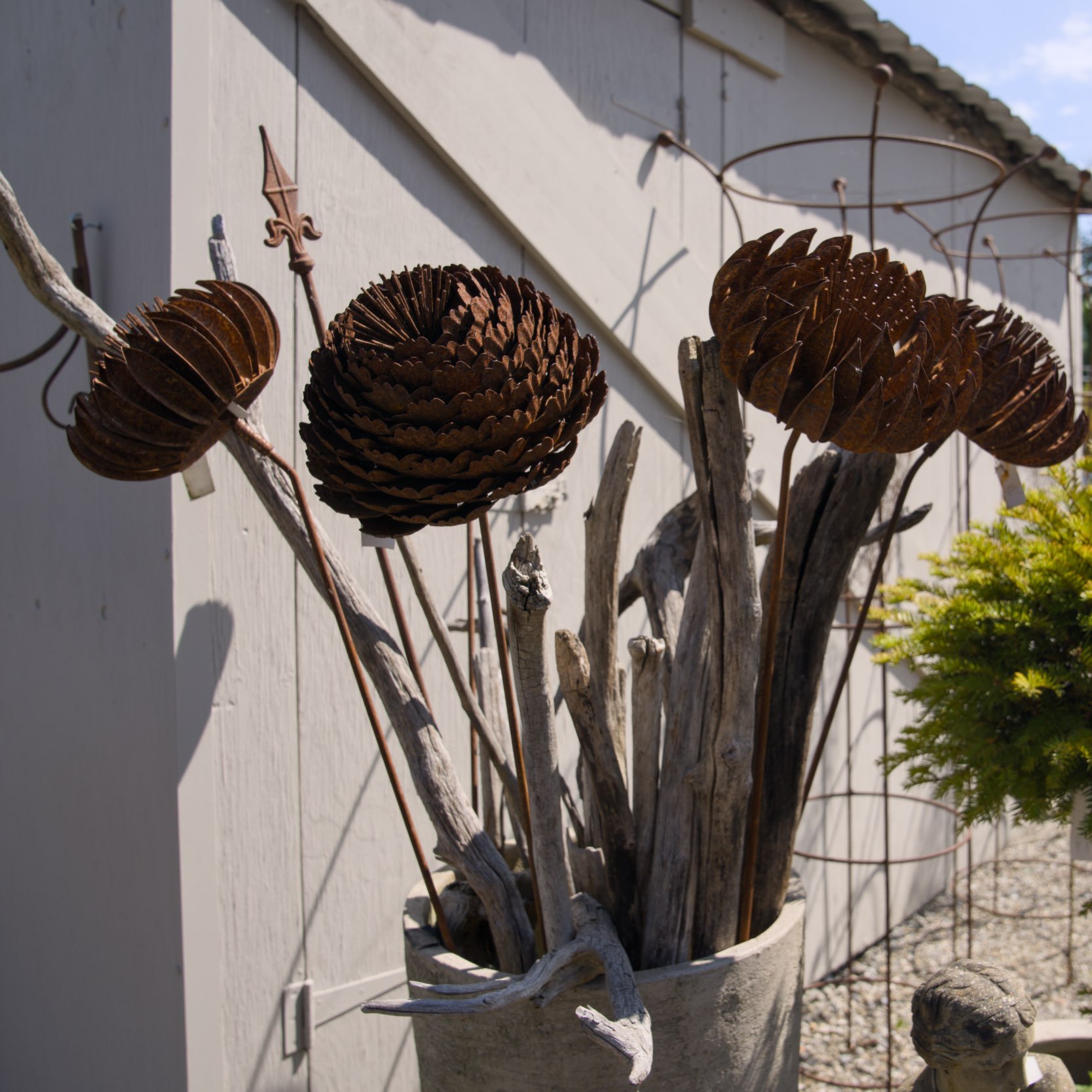 Kunstvolle Metallskulptur, gestaltet wie große, spiralförmige Blüten oder Früchte, an einem weißen Gartenzaun mit Holzstäben und Draht.