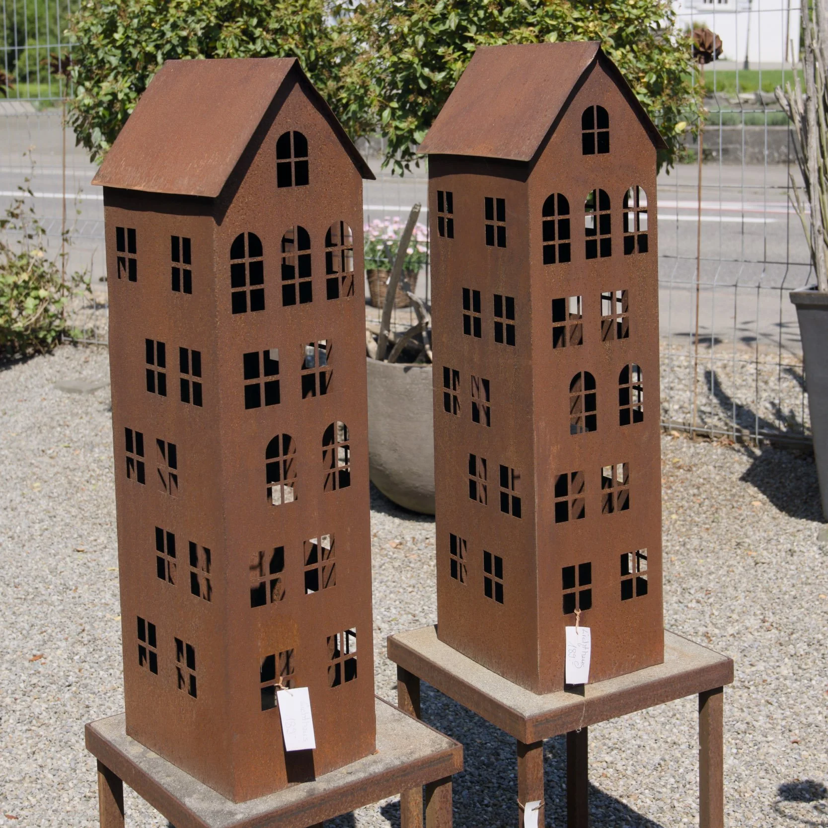 Zwei metallene Miniaturhäuser auf Holzständern, mit mehreren Fenstern und Dächern, im Freien.