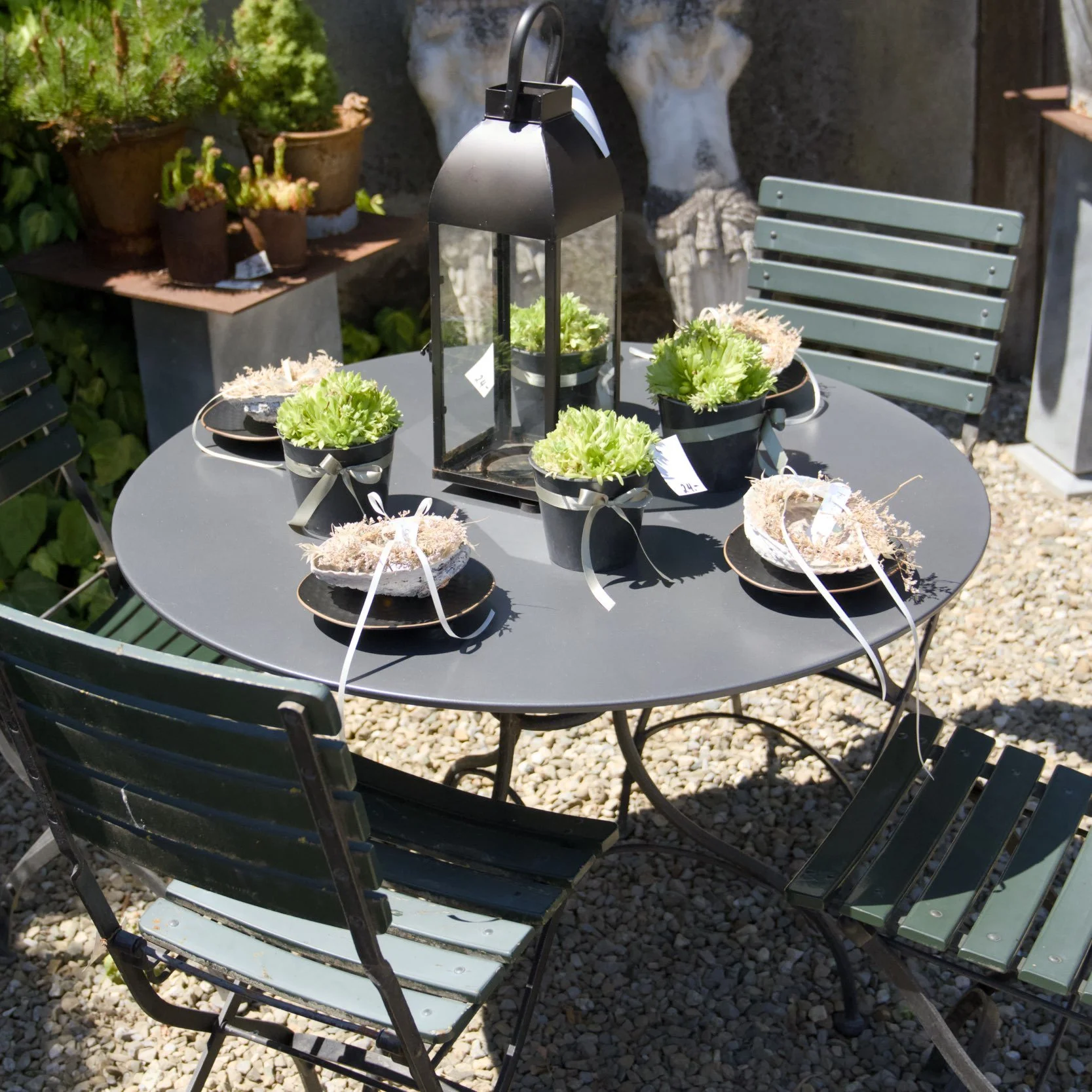 Ein runder Gartentisch mit sechs schwarzen Blumentöpfen und einer dekorativen Laterne. Der Tisch steht auf einem Kiesboden im Freien, im Hintergrund Pflanzen und eine Skulptur aus Stein.