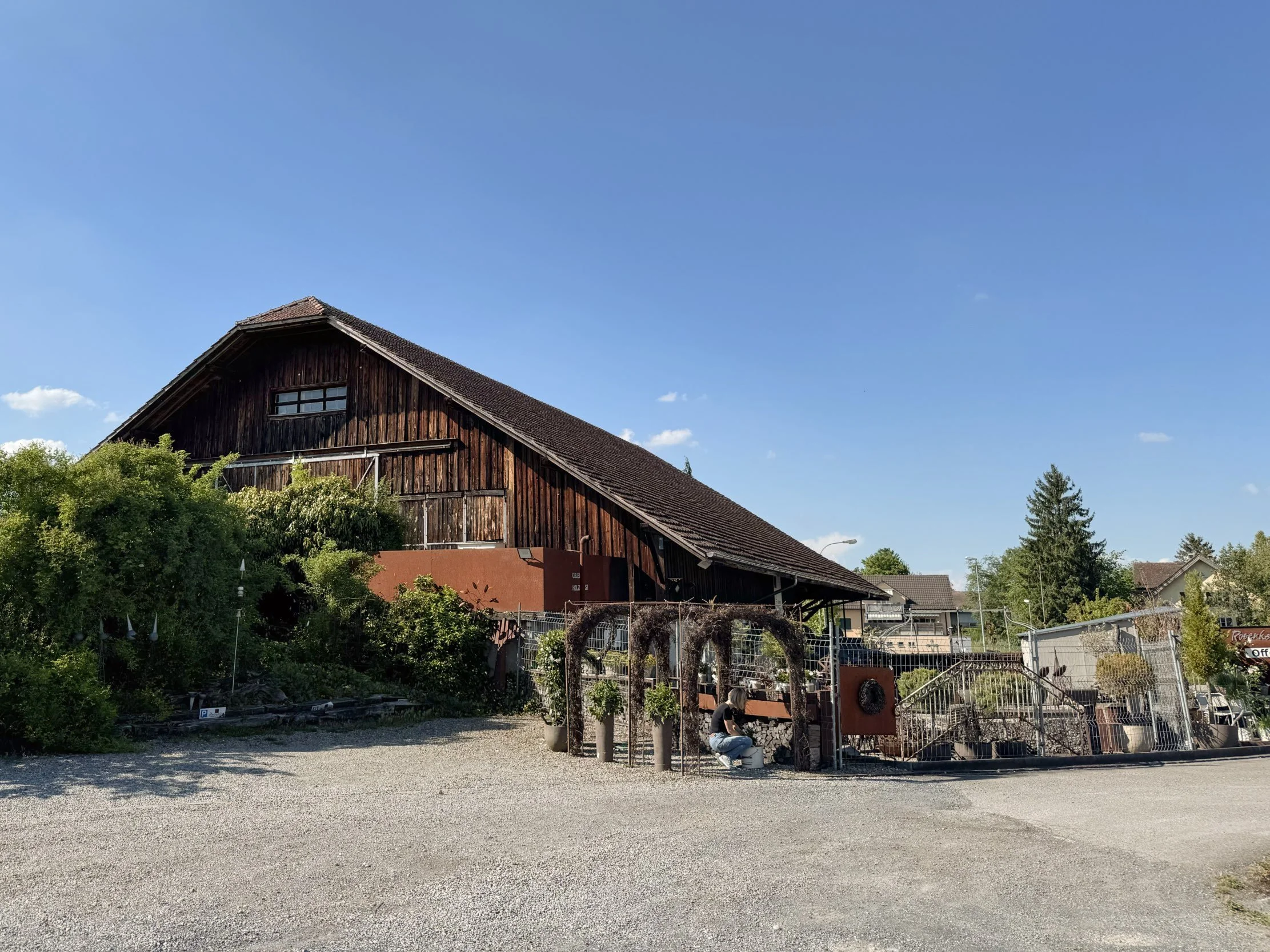Ein landhausartiges Gebäude mit einer schrägen Holzfassade und einem Dach. Im Vordergrund befindet sich ein geräumiger Parkplatz mit einem Menschen, der an einem kleinen Gemüsegarten arbeitet, umgeben von Bäumen und Gebüschen. Der Himmel ist blau mit