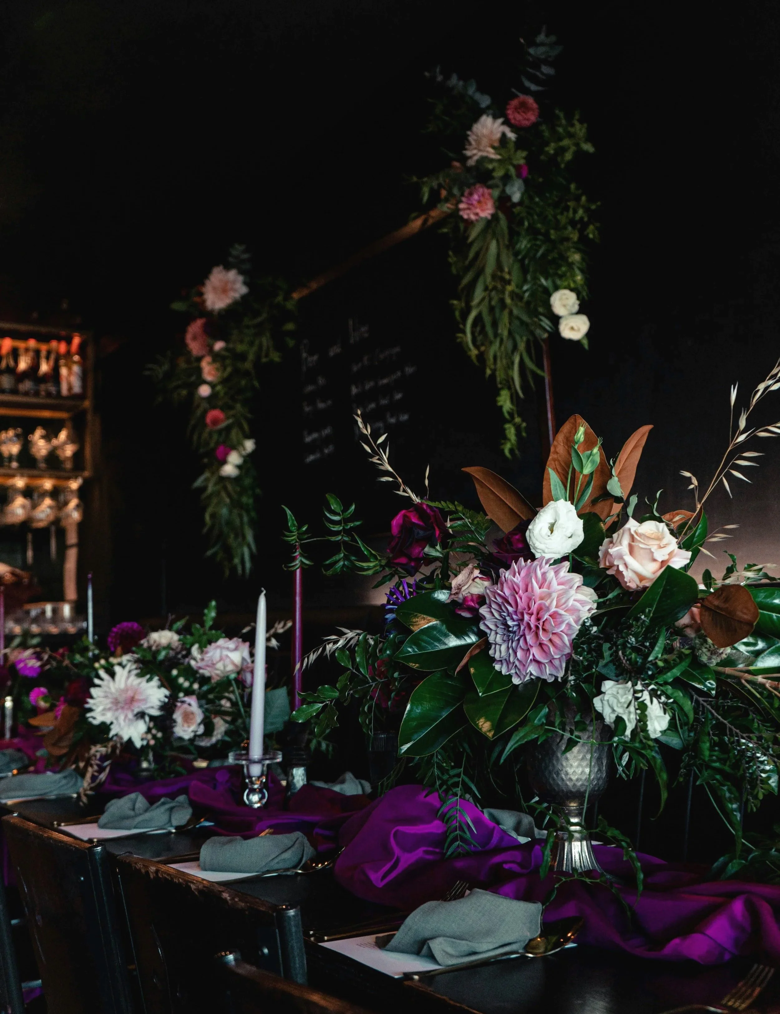 Blumenarrangement mit weißen, rosa und violetten Blumen in einer silbernen Vase, dekoriert mit grünen Blättern und braunen Zweigen, auf einem Tisch mit Stoffdecken in Violett und Grau, im Hintergrund eine schwarze Wand mit einem Blumenkranz und einer