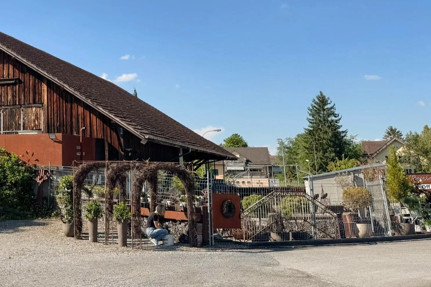 Ein ländliches Gebäude mit Holzwand, davor ein Zaun und eine Frau, die einen Eimer hält, in einer ländlichen Umgebung, mit Bäumen und Häusern im Hintergrund, bei sonnigem Wetter.