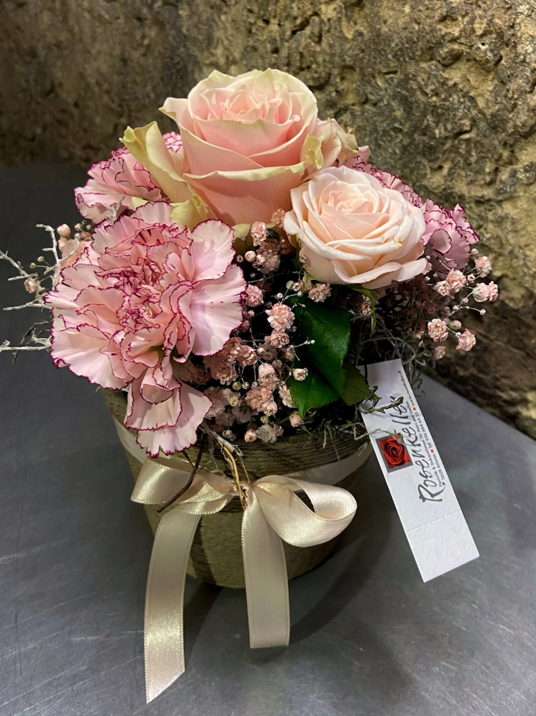 Ein Blumenstrauß mit zwei großen, cremefarbenen Rosen, pinkfarbenen Hortensien mit roten Rändern, kleinen rosa Blumen und Gräsern, in einem Korb mit goldener Schleife, auf einer dunklen Oberfläche vor einer Steinmauer.