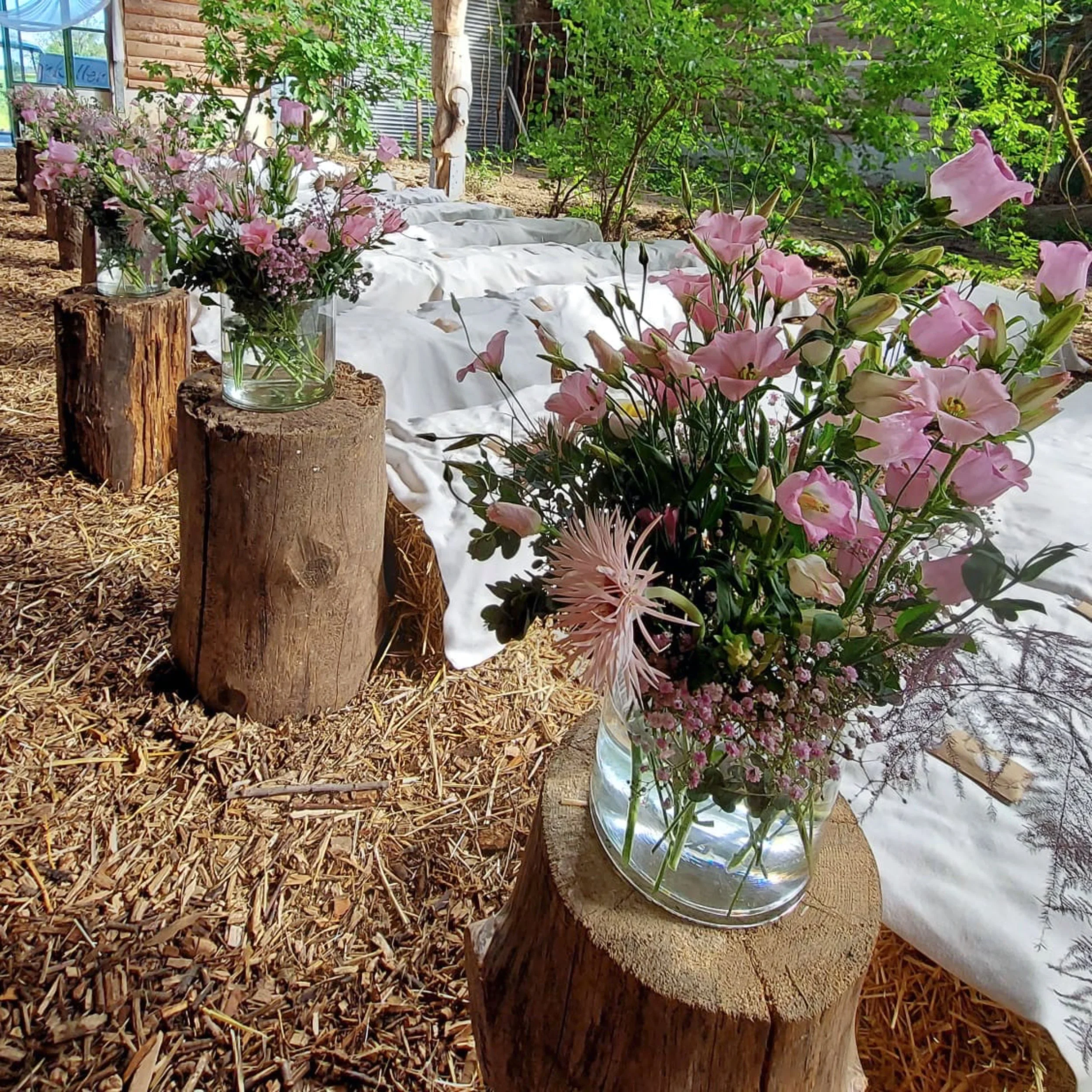 Bunte Blumenarrangements in Gläsern, umgeben von Baumstämmen und Holzspänen, in einer grünen Gartenumgebung.