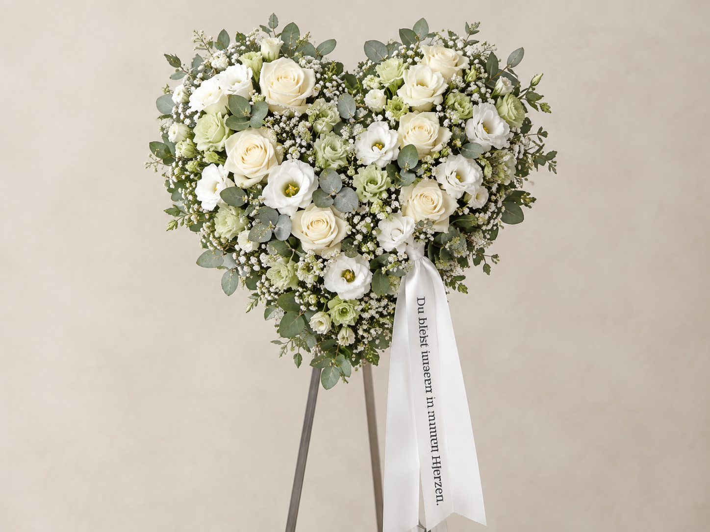 Blumenkranz in Herzform aus weißen Rosen, Eustoma, Schleierkraut und Eukalyptus, mit weißer Schleife mit deutschem Trauerspruch auf tristem Hintergrund.