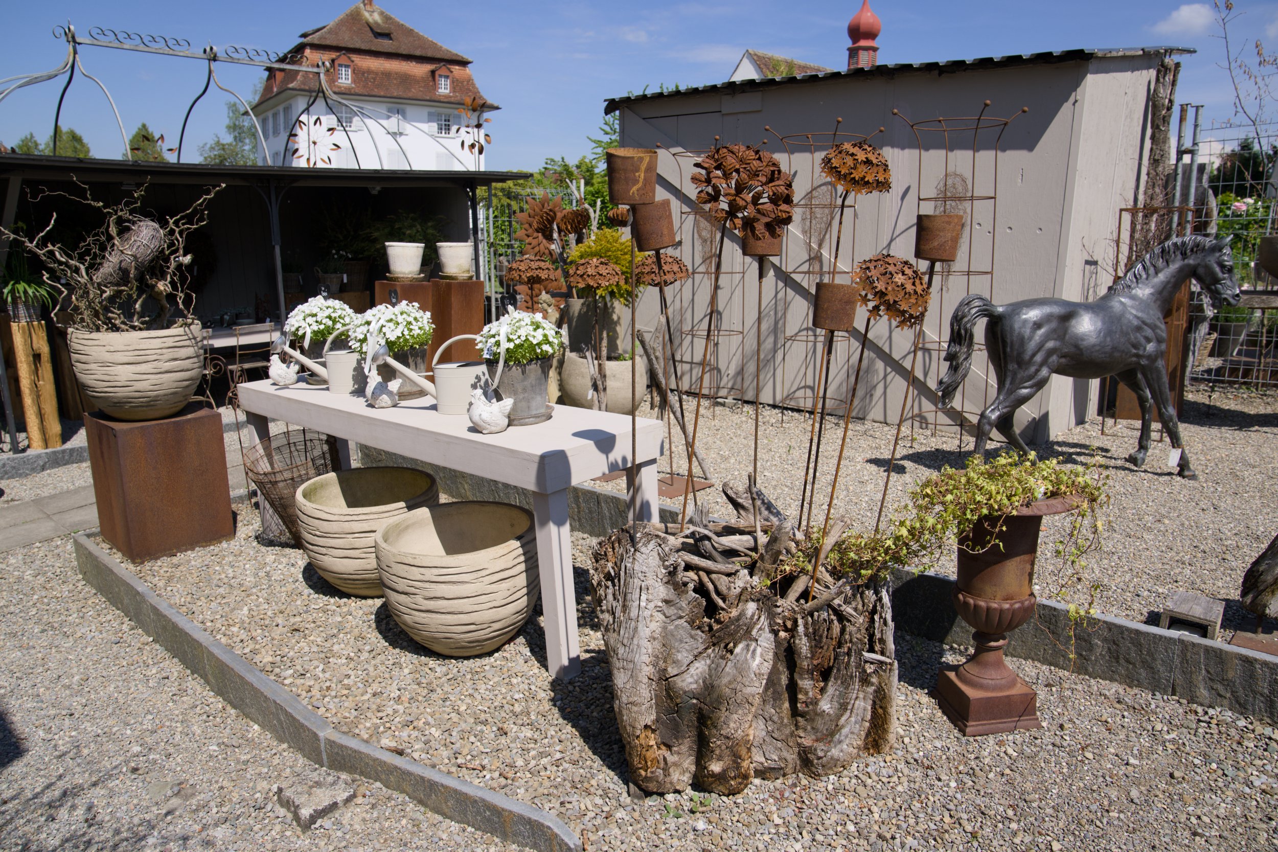 Garten mit Skulpturen, Blumen und Dekorationen, im Hintergrund ein alter Holzzaun und ein Haus mit roten Ziegeln. Es gibt einen weißen Tisch mit weißen Blumen und Gartendekorationen, sowie verschiedene metallene und steinerne Kunstwerke, darunter ein