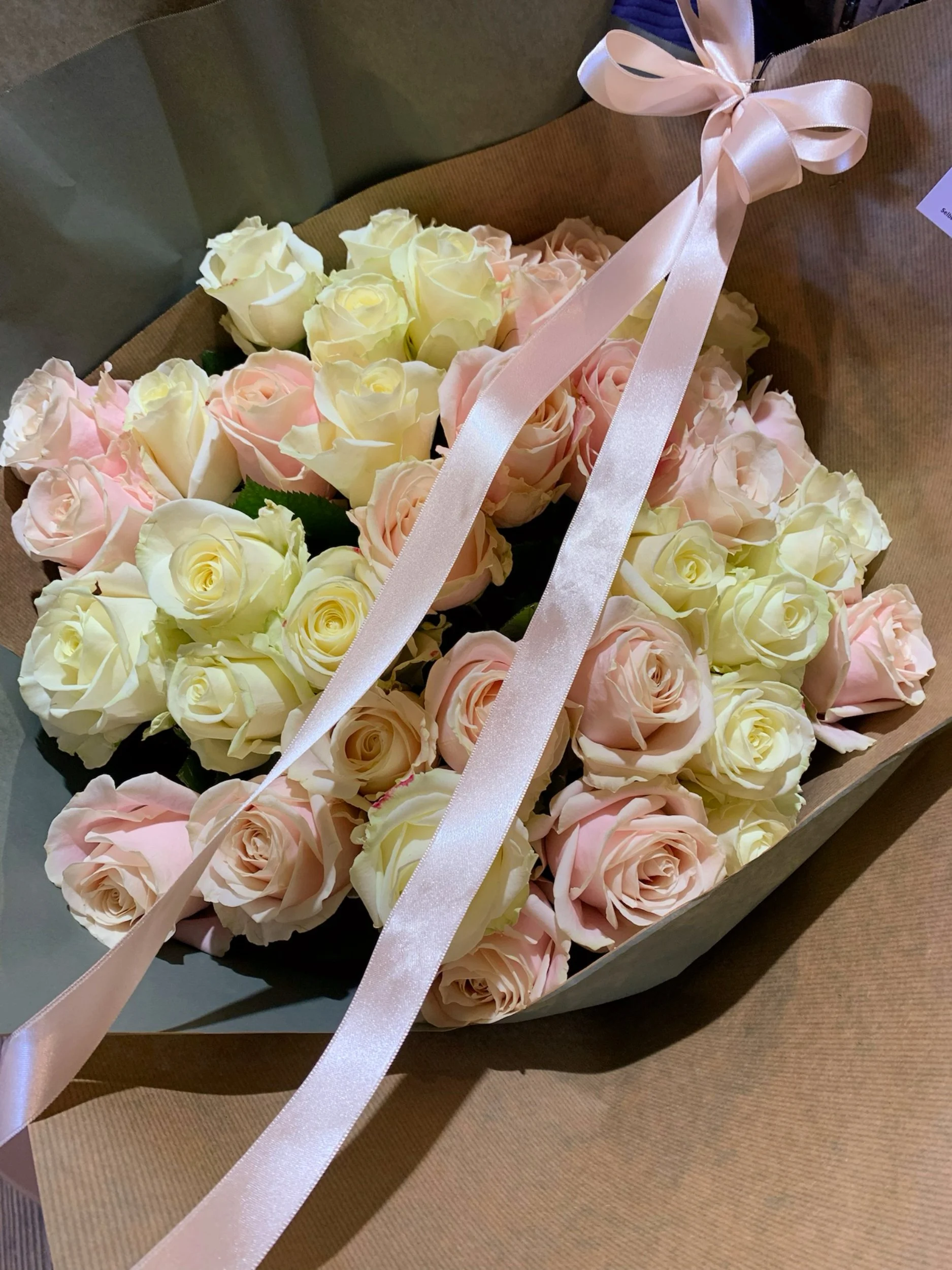 Ein Blumenstrauß aus weißen und rosa Rosen, dekoriert mit einem zarten rosa Band, liegt in einer Schachtel auf braunem Papier.