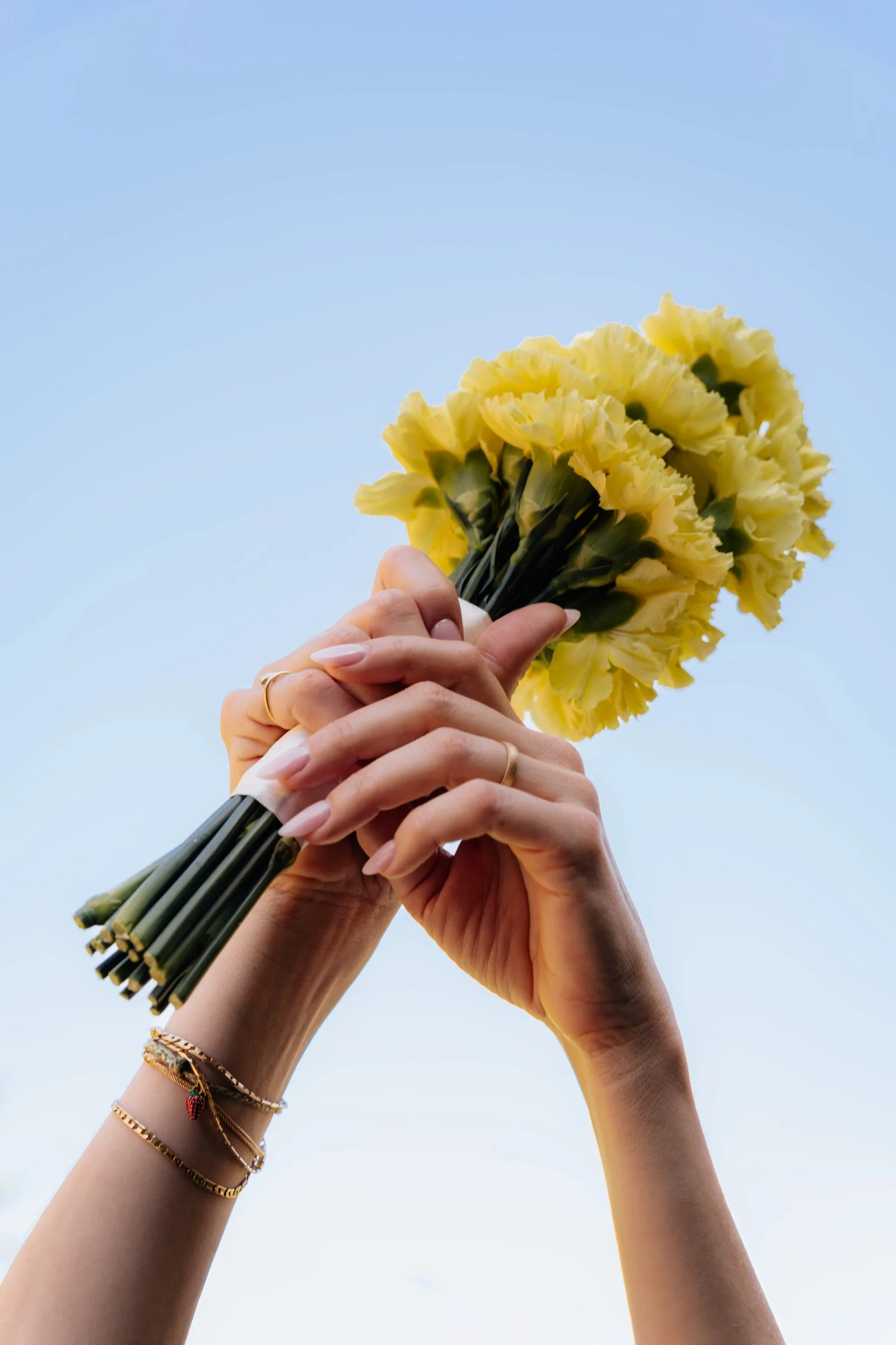 Zwei Hände halten einen Bund gelber Blumen gegen den Himmel. Die Hände tragen mehrere goldene Armbänder.