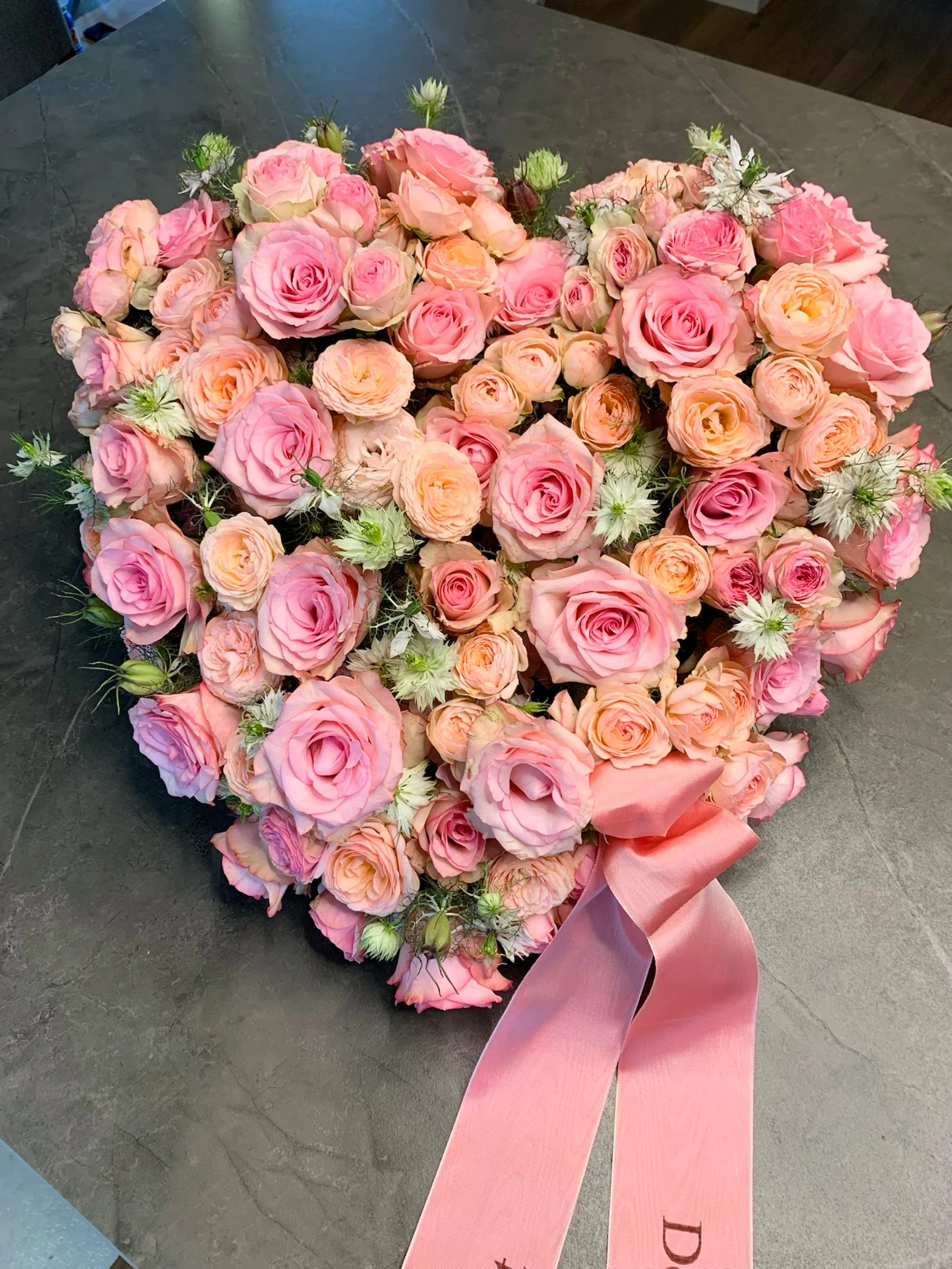 Ein Herz aus rosa und peachfarbenen Rosen, mit weißen Blüten und grünen Blättern, verziert mit einer großen rosa Schleife, liegt auf einem dunklen Steinboden.