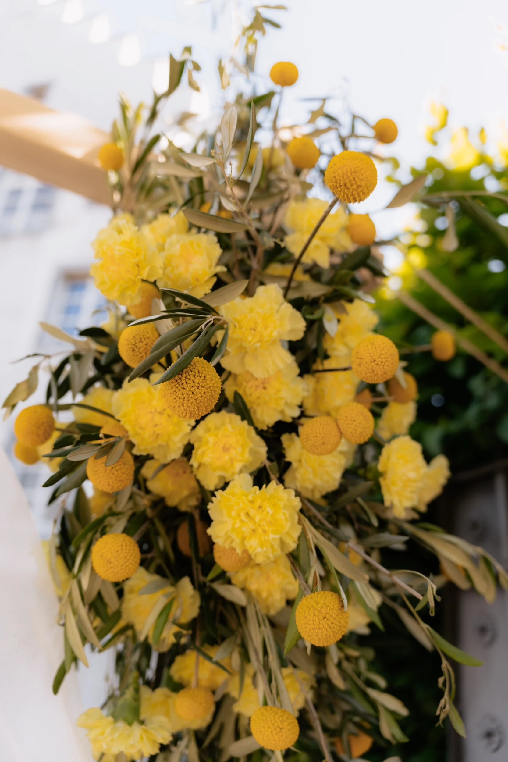 Blumenstrauß mit gelben Blüten, darunter Olivenzweige. Hintergrund hell und unscharf.