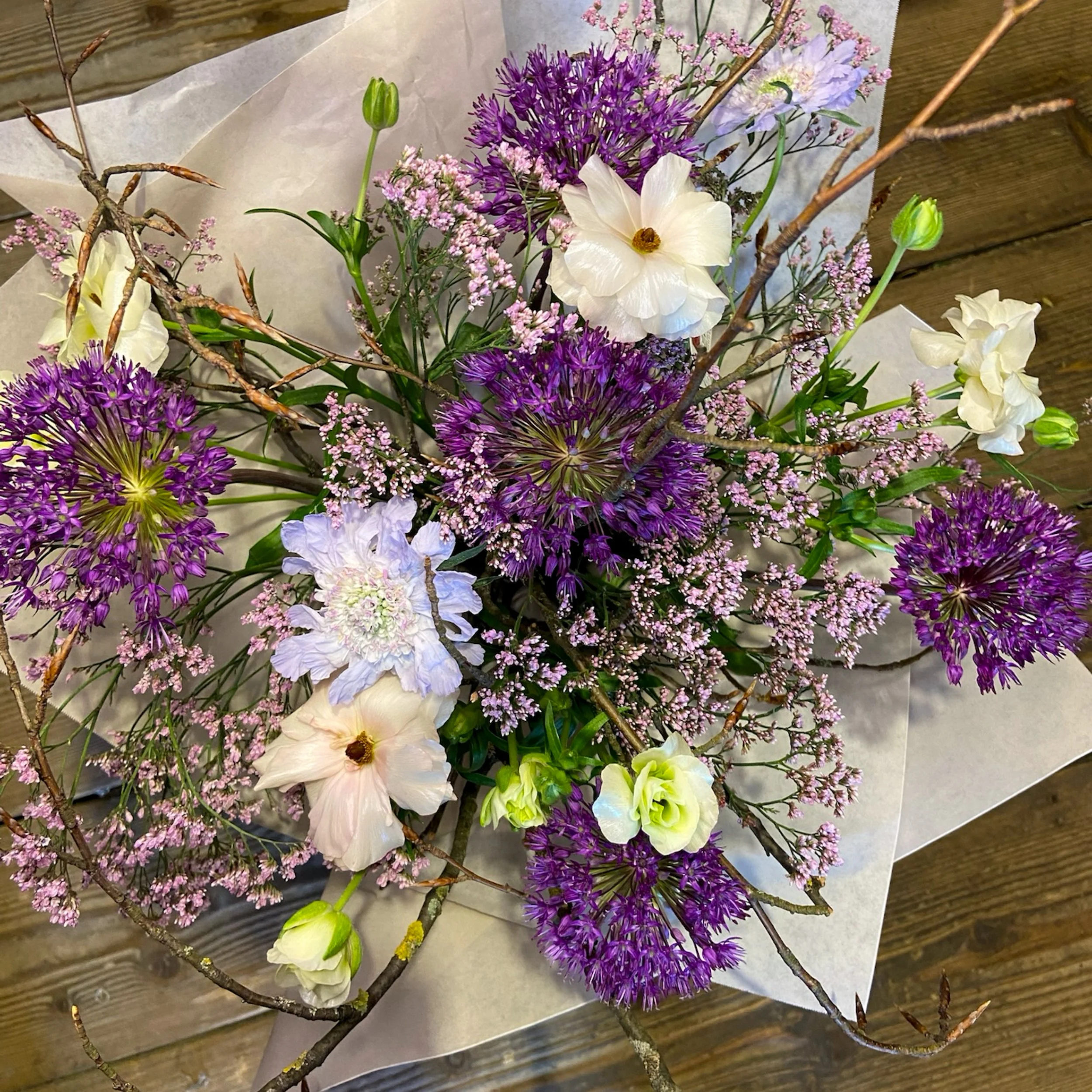 Blumenstrauß mit weißen, lila, pinken Blumen und grünen Knospen, verziert mit gebrochenen Ästen, auf einem Holztisch.