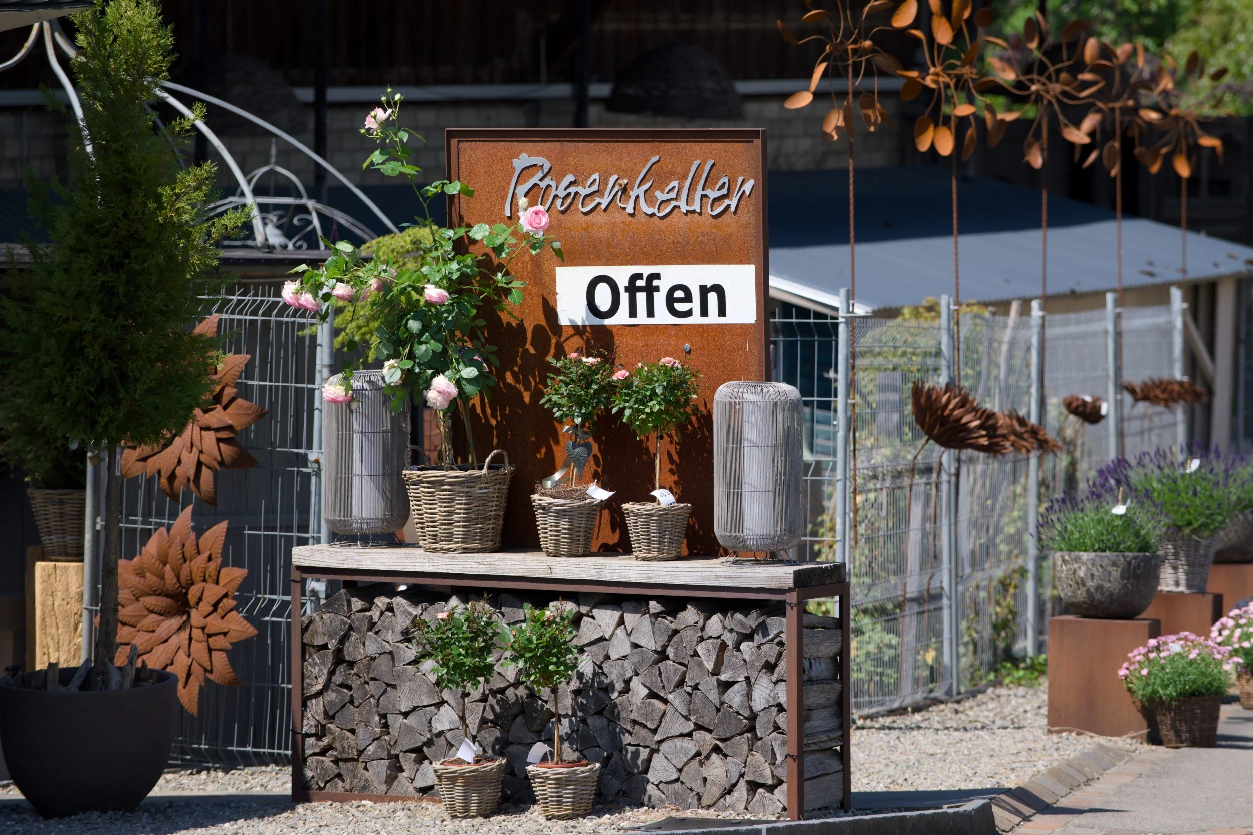 Ein Gartenschild mit der Aufschrift "Offen" steht vor einem Regal mit Blumen und Holzscheiten im Hintergrund.