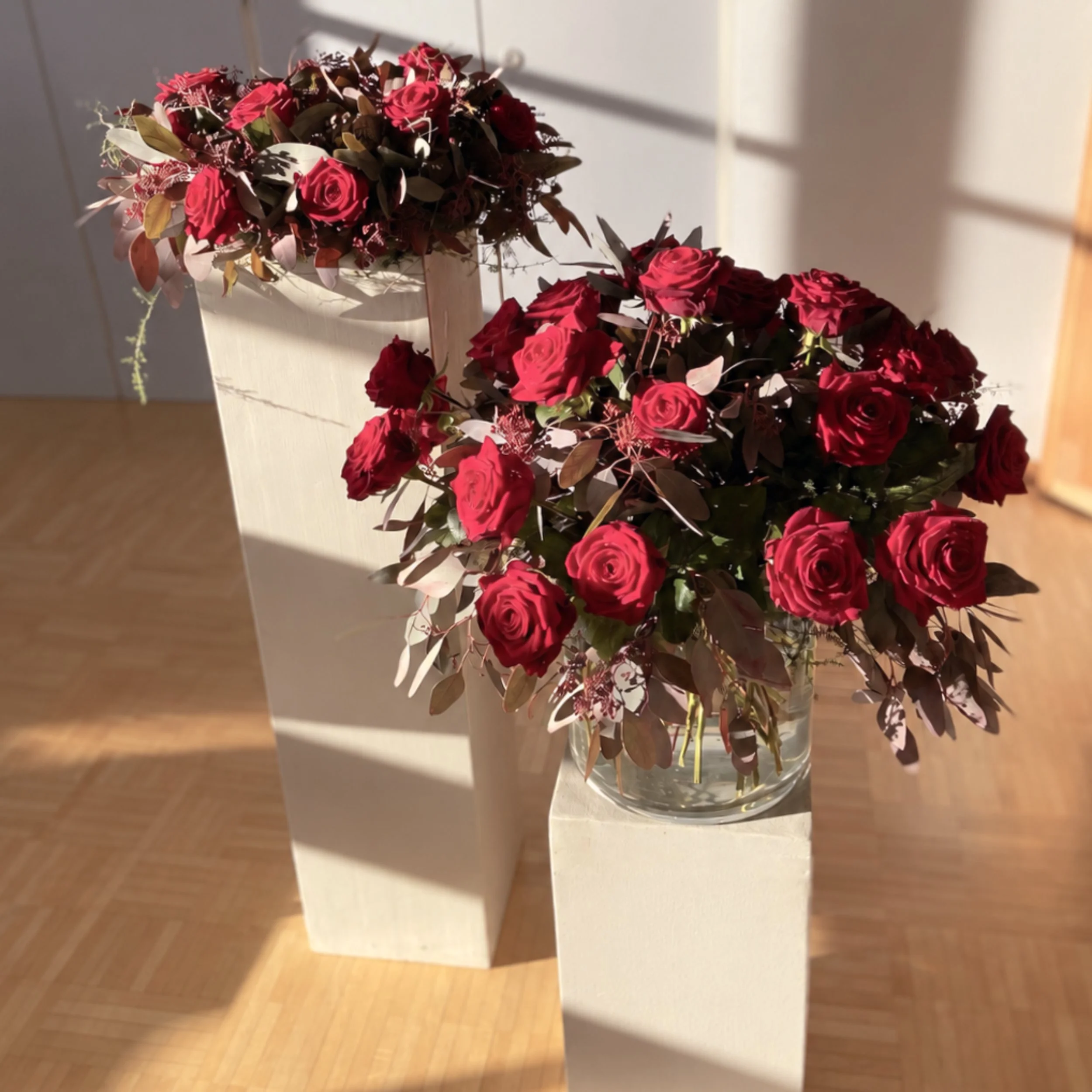 Blumenstrauß mit roten Rosen und grünem Laub in einer Vase auf einem weißen Tisch, im Hintergrund eine graue Wand.