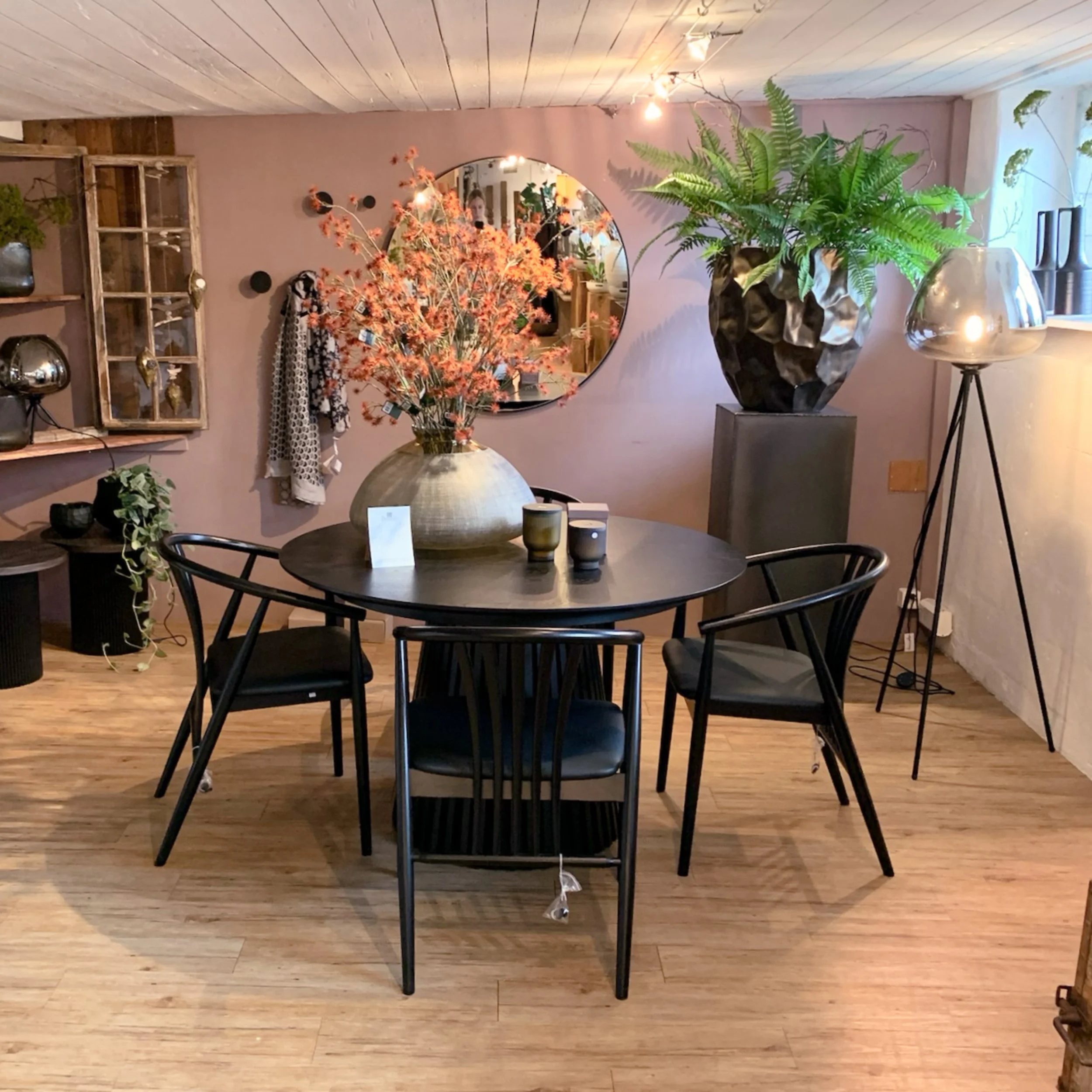 Ein Blick auf einen modernen Innenraum mit einem runden Esstisch aus dunklem Holz, umgeben von schwarzen Stühlen. Auf dem Tisch steht eine große Vase mit pinkfarbenen Blumen. Im Hintergrund befinden sich eine Wand mit einem runden Spiegel, einige Dek