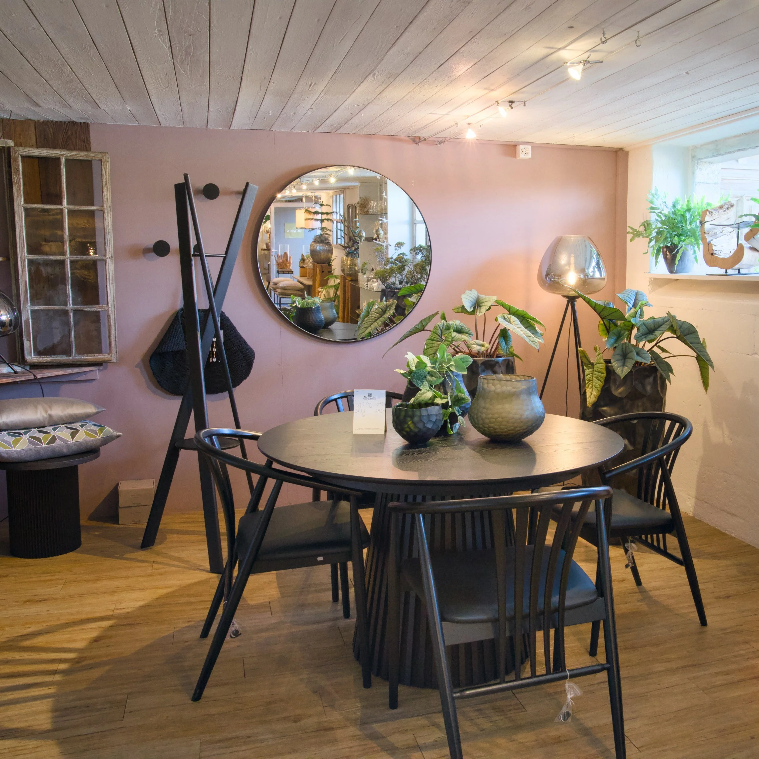 Ein Wohnzimmer mit pastellrosa Wand, runden Spiegel, Pflanzen in Vasen, Lampen und ein Esstisch mit Stühlen