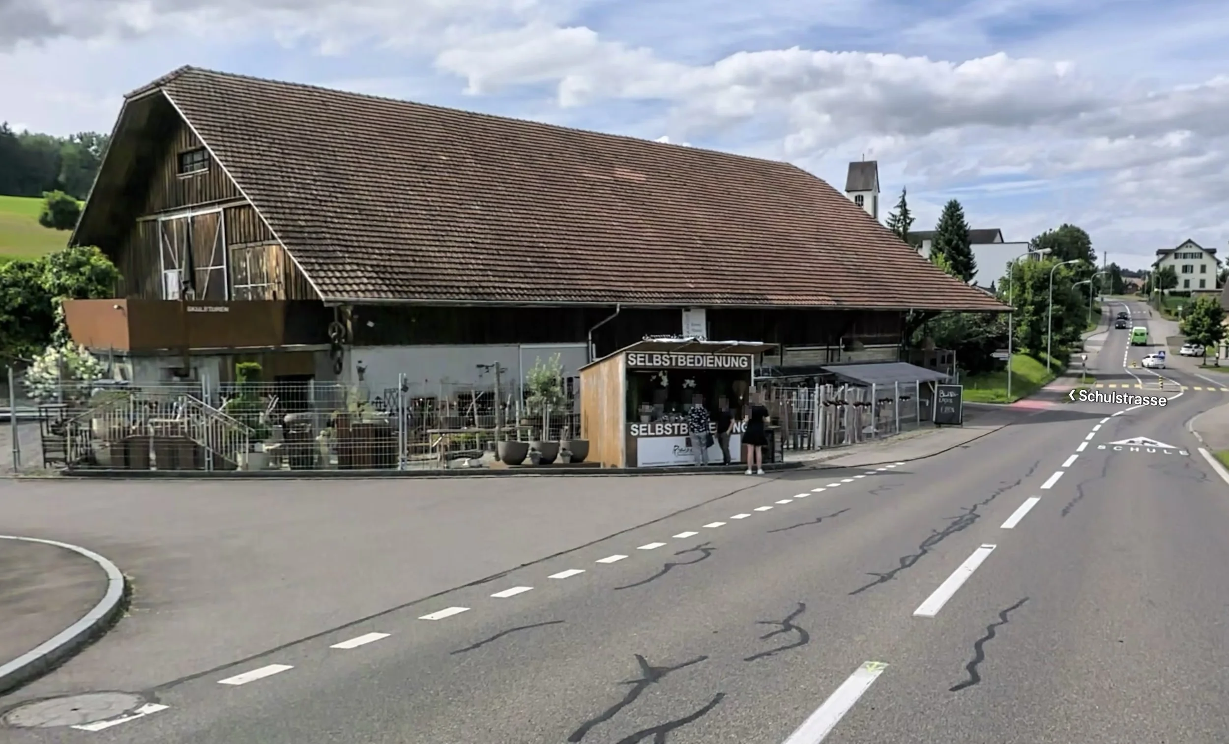 Ein großes, schräg geneigtes Holzgebäude an einer Straße mit einem Selbstbedienungsladen davor, auf einer Seite befindet sich eine Terrasse mit Pflanzen. Im Hintergrund sind Häuser und Bäume sichtbar, der Himmel ist bewölkt.
