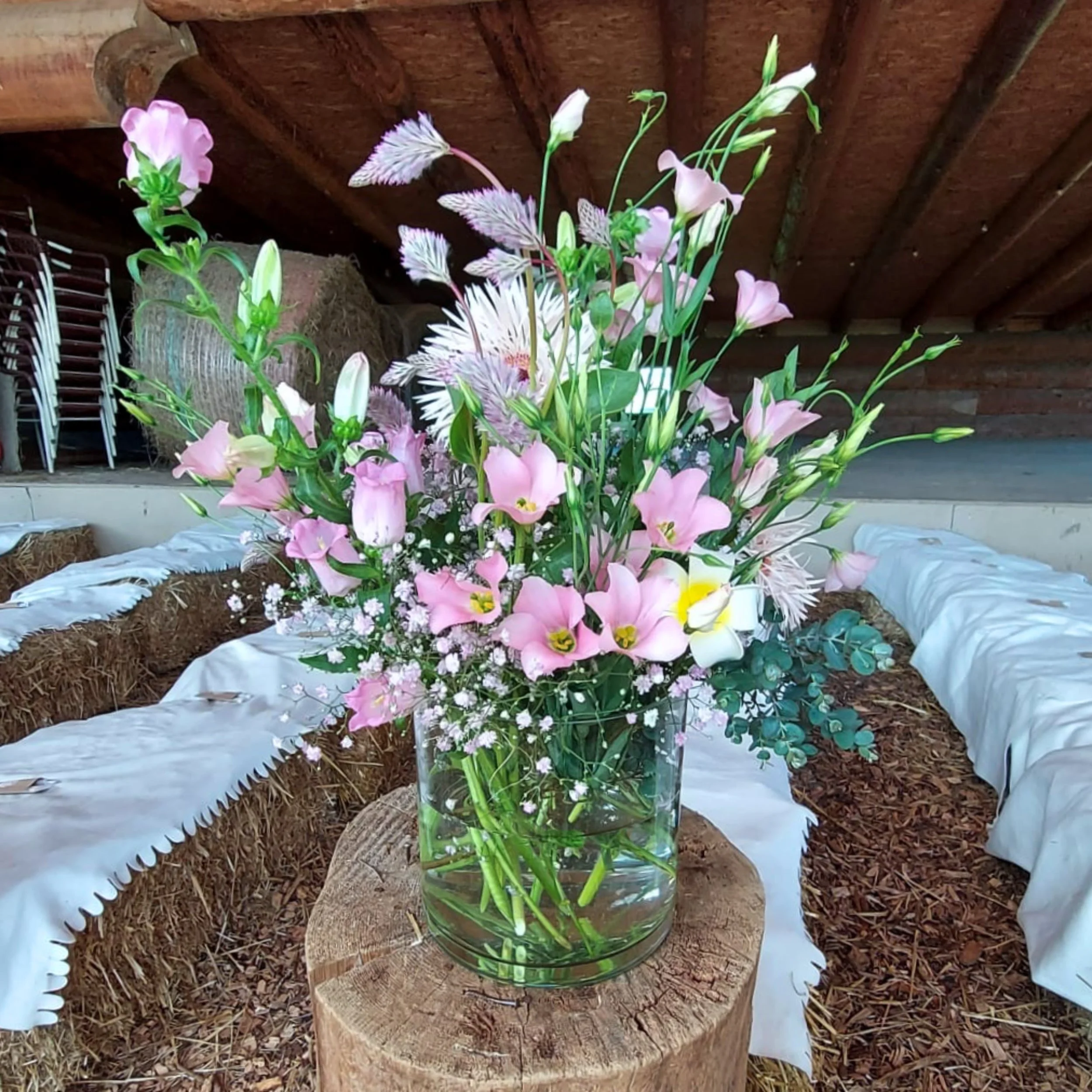 Blumenstrauß mit rosa und weißen Blumen in einer Glaskaraffe auf einem Baumstumpf, im Hintergrund Holzboden und Stuhl.