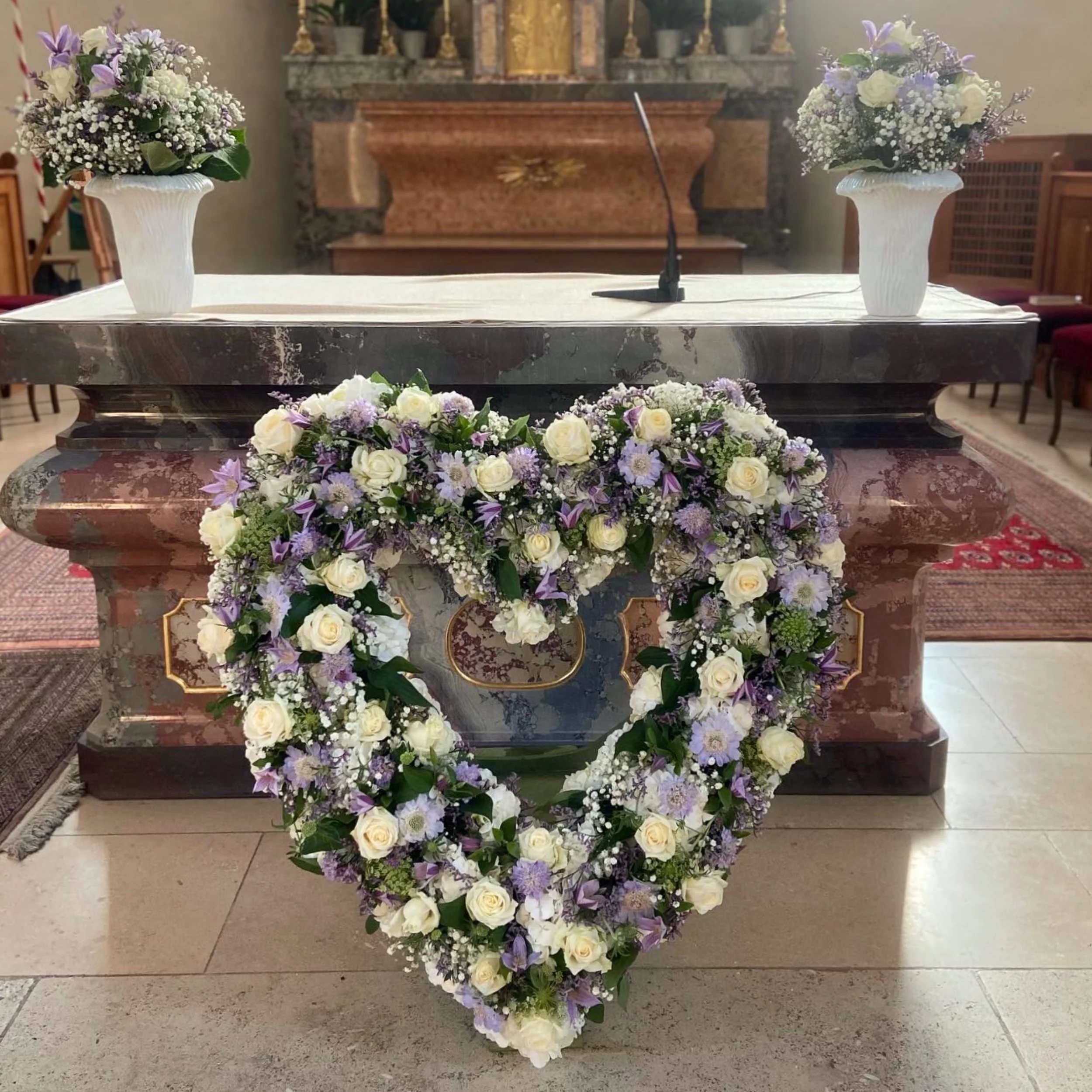 Blumenarrangement mit weißen Rosen und lila und weißen kleineren Blumen, dekorativ auf einem Altar in einer Kirche platziert.