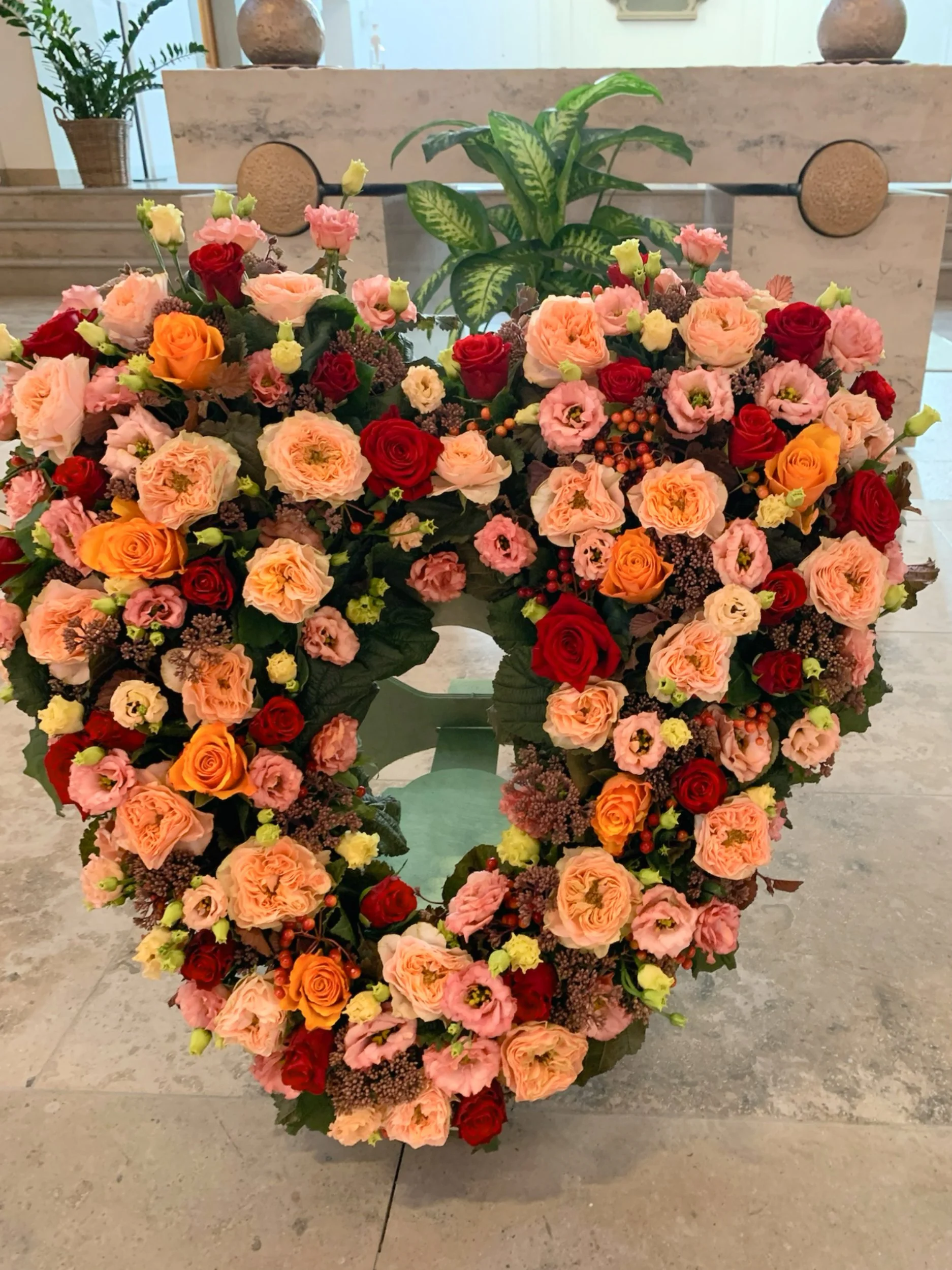 Großes Blumenarrangement mit roten, rosa, orange und gelben Rosen, ergänzt durch lilafarbene und grüne Pflanzen, vor einem weißen Marmorblock, im Hintergrund Pflanzen und Dekoration.