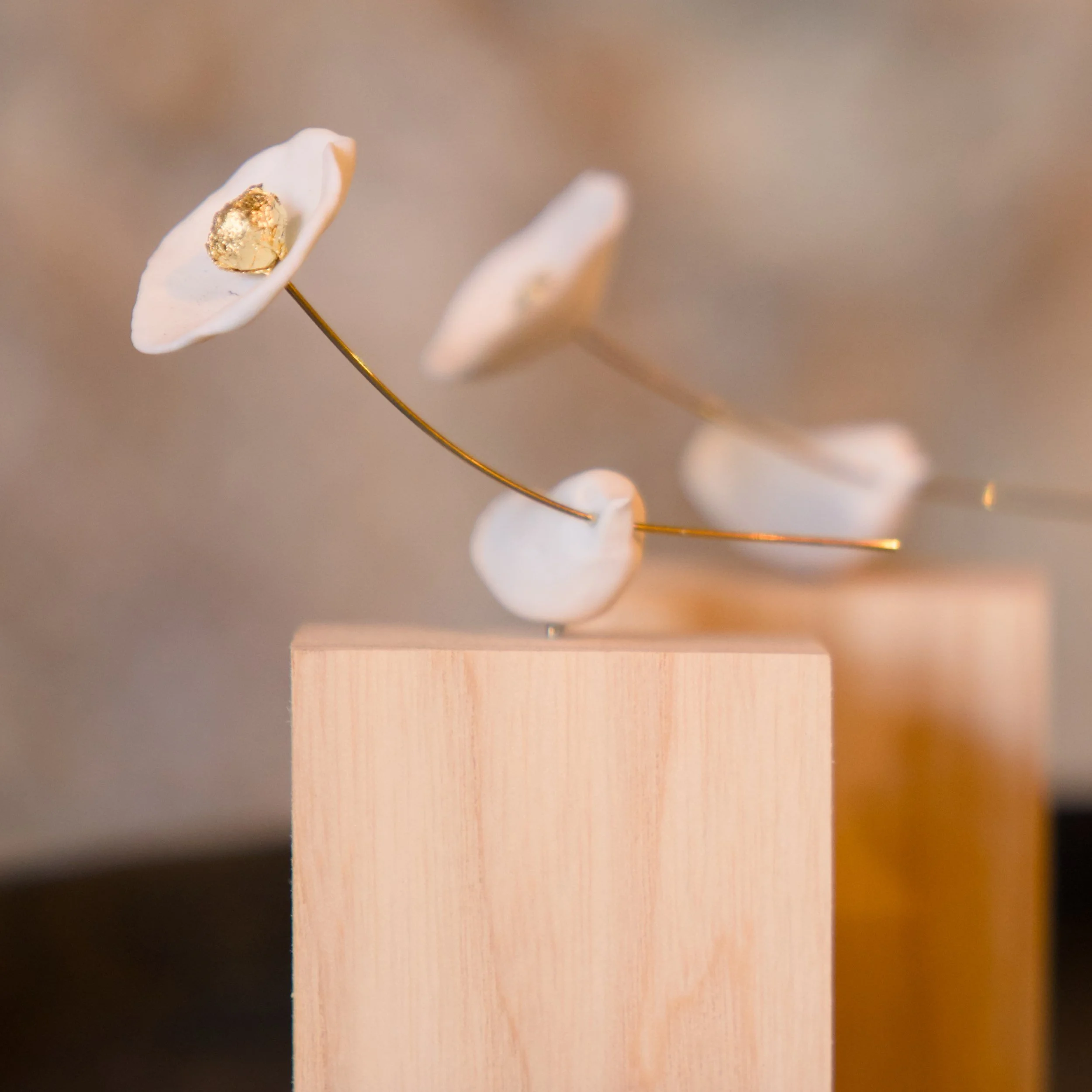 Eine dekorative Skulptur, bestehend aus weißen Blättern oder Blüten mit goldenen Akzenten auf dünnen, goldfarbenen Stäben, steht auf einem rechteckigen Holzteller. Der Hintergrund ist unscharf und in neutralen Farben gehalten.
