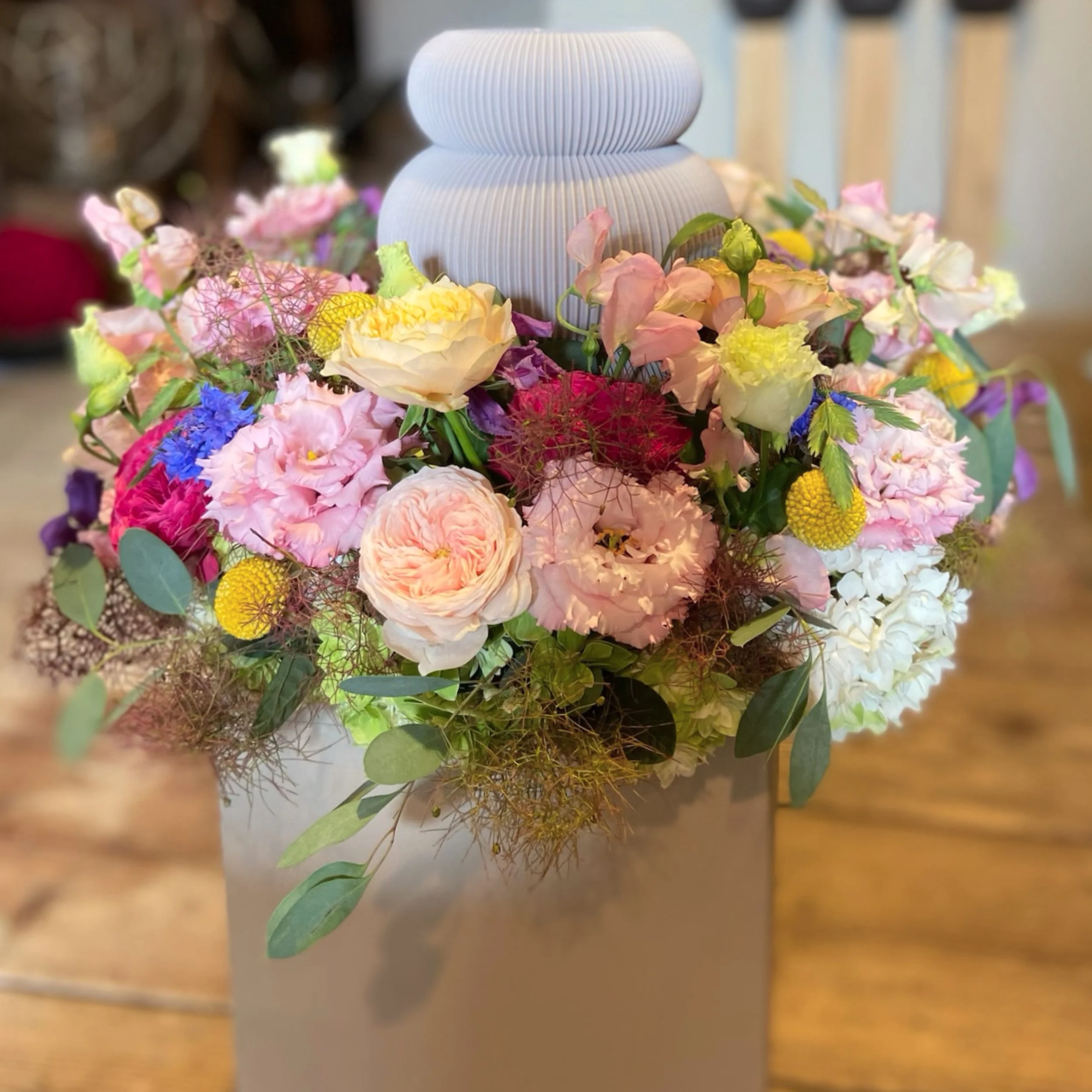 Vase auf einem Tisch, umgeben von bunten Blumen in verschiedenen Farben wie rosa, gelb, lila, und grün, mit grünem Laub.