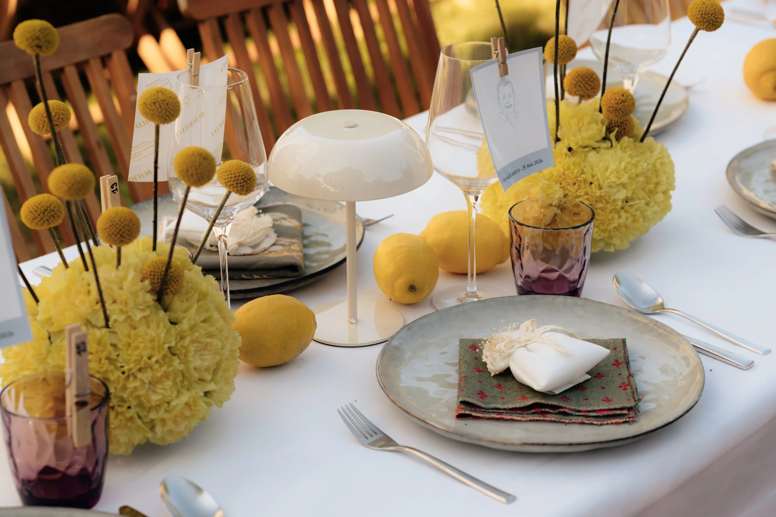 Ein festlich gedeckter Tisch mit gelben Blumen, Zitronen, leeren Weingläsern, einem weißen Kerzenleuchter und Teller mit Besteck, bereit für eine Mahlzeit im Außenbereich bei Sonnenlicht.