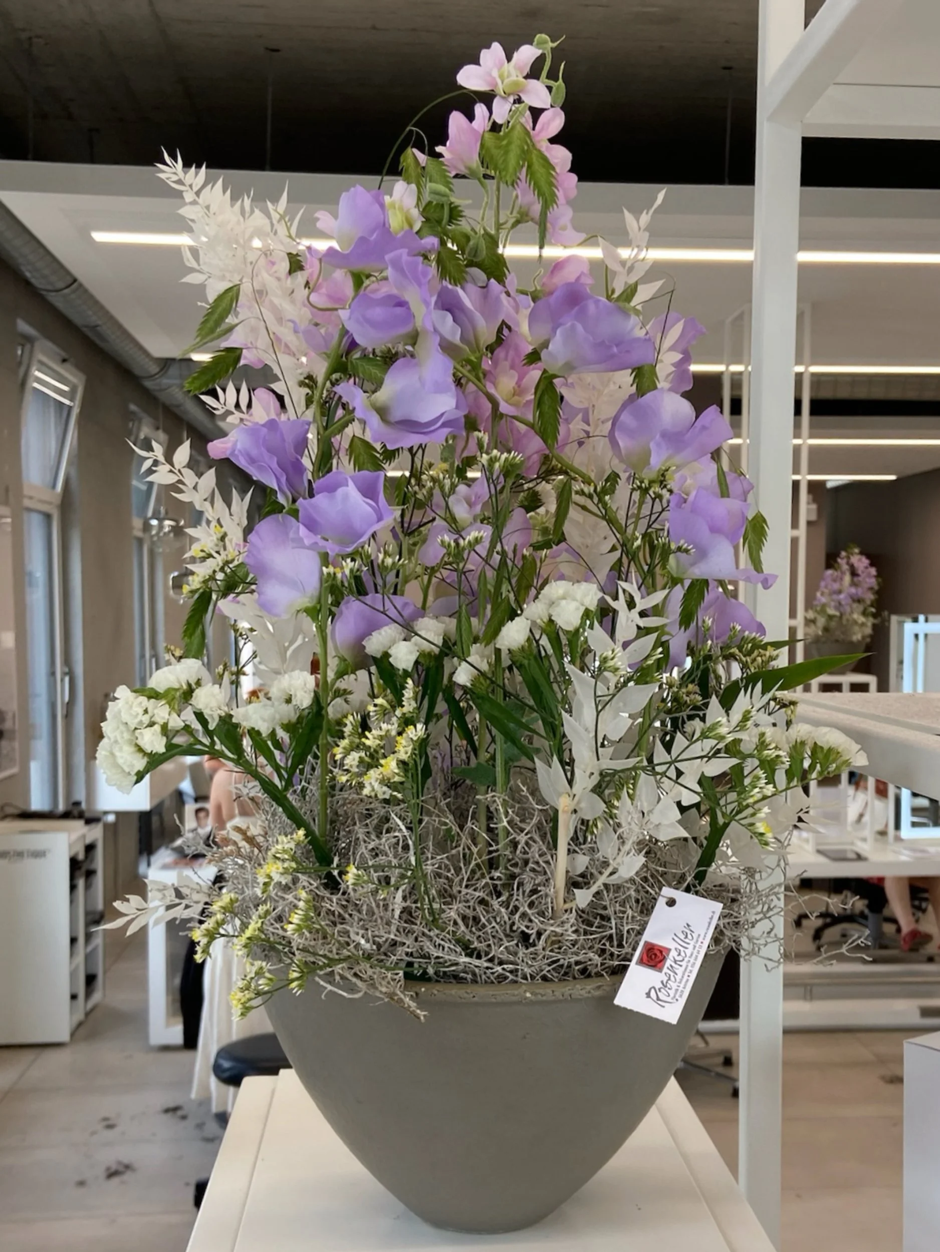 Blumenarrangement mit weißen, lilafarbenen und grünen Blumen in einer großen grauen Vase auf einem weißen Tisch im Raum.
