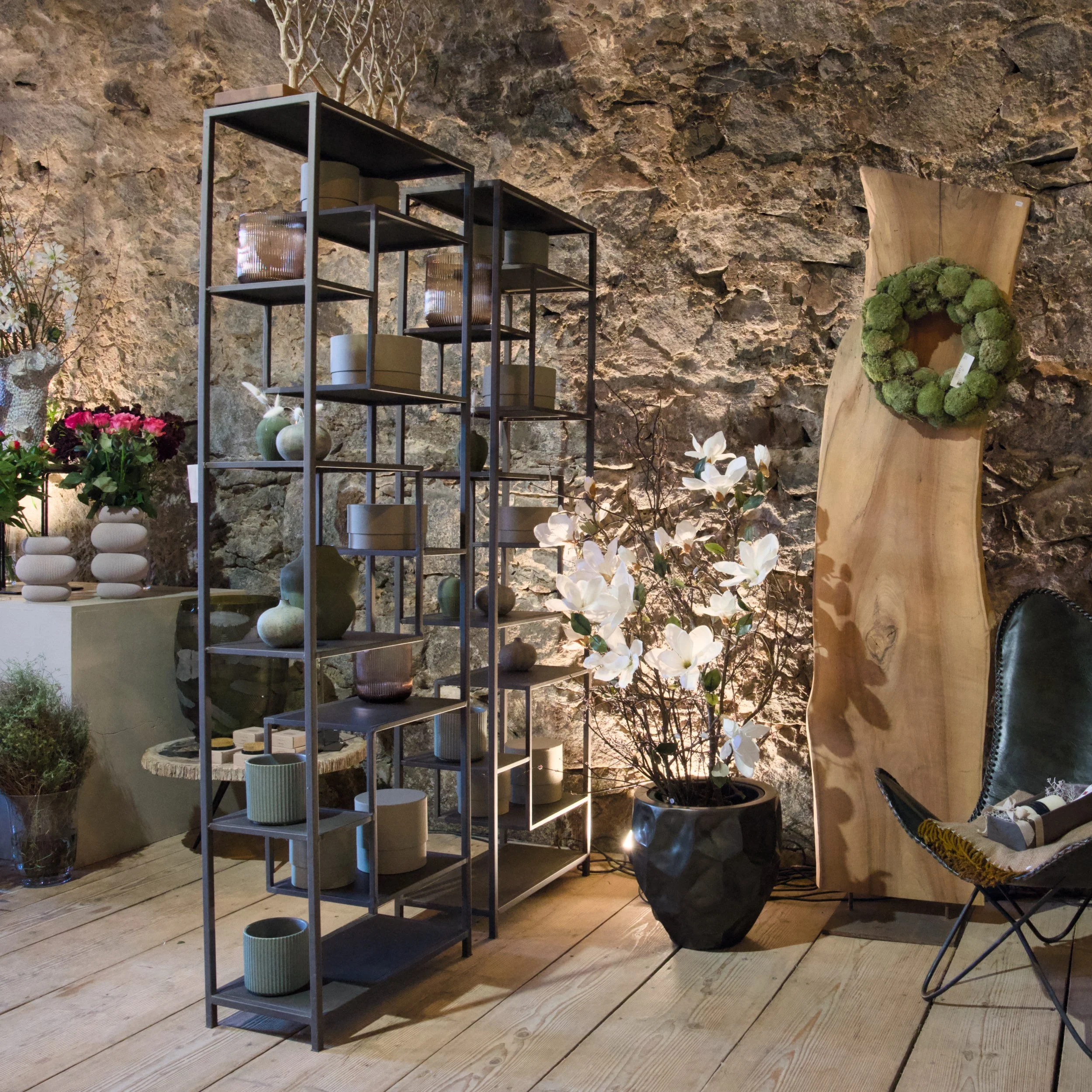 Innenraum eines stilvollen Ladens oder Showrooms mit einer Stehliste aus Metall, die Keramiktöpfe enthält. Im Hintergrund eine Wand aus rohem Stein, umliegende Pflanzen und dekorative Gegenstände auf Holzboden.