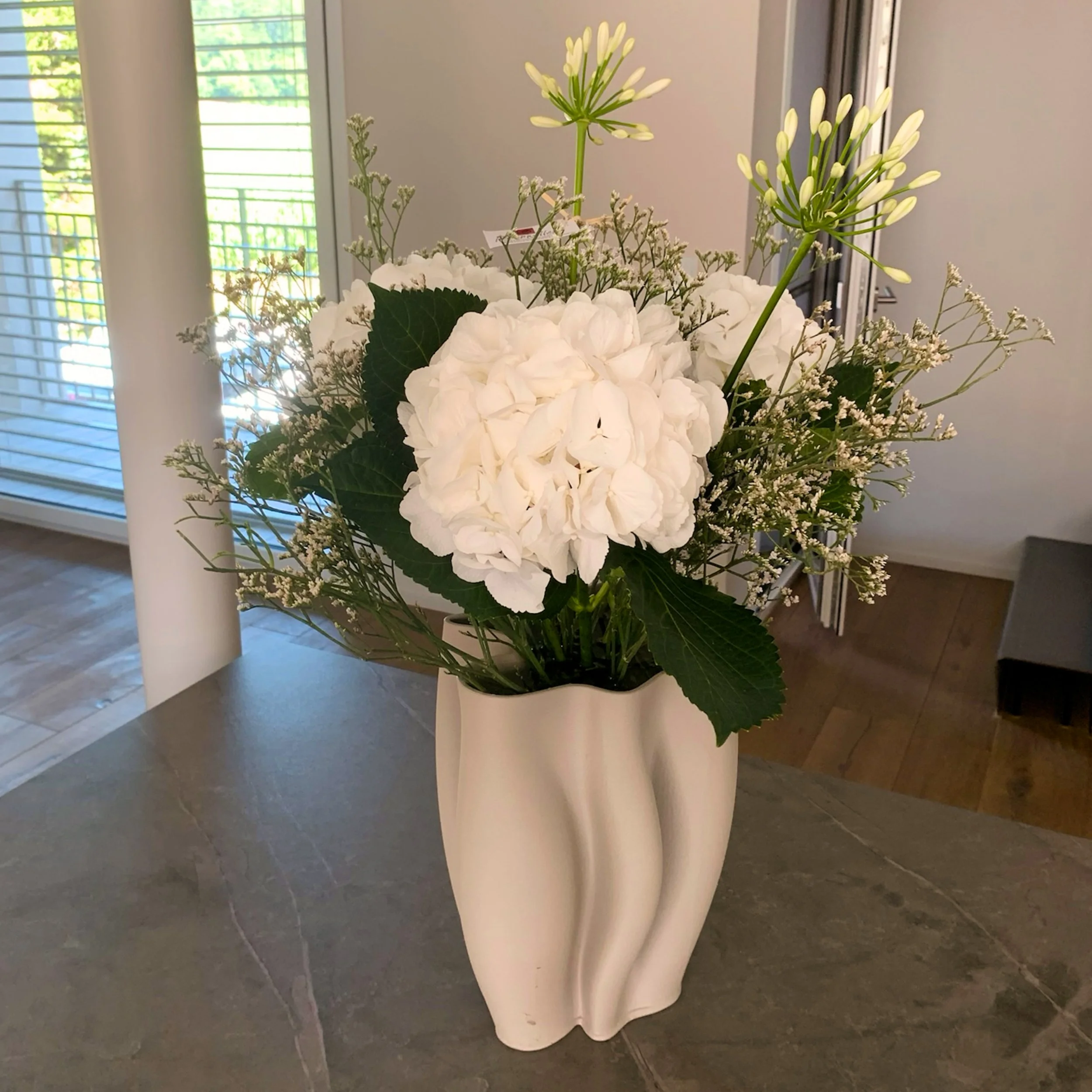 Eine weiße Vase mit einer Anordnung von weißen Hortensien, kleinen weißen Blumen und weißen Lilien, auf einem weißen Tisch in einem Raum mit Fenster und Rollläden im Hintergrund.