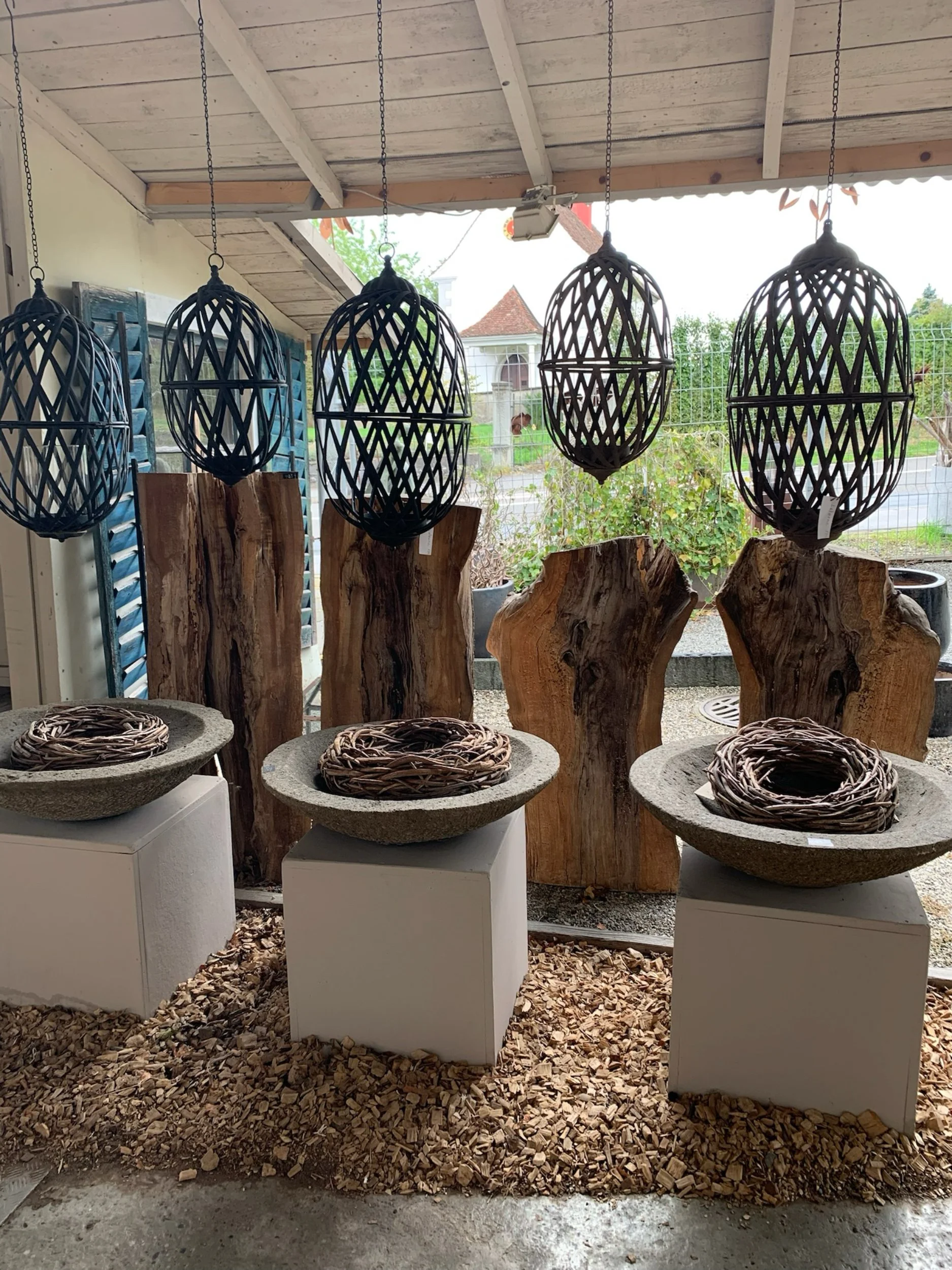 Vier schwarze, gitterartige Laternen hängen an einer Holzdecke, im Hintergrund ein Garten mit einer Gartenmauer und einem Haus, im Vordergrund befinden sich drei große, runde Sitzschalen mit Sitzkörben auf weißen Säulen, auf dem Boden liegt Mulch.