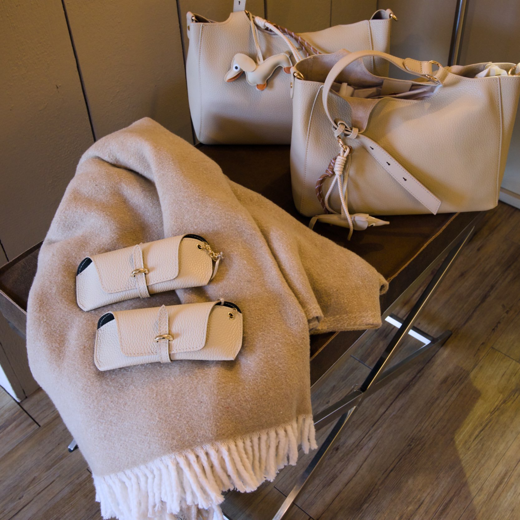 Auf einem Tisch liegen drei beige Handtaschen, eine mit einem Schlüsselanhänger in Form einer Ente. Zudem liegt ein beige Mantel und einige kleinere Gegenstände auf dem Tisch.