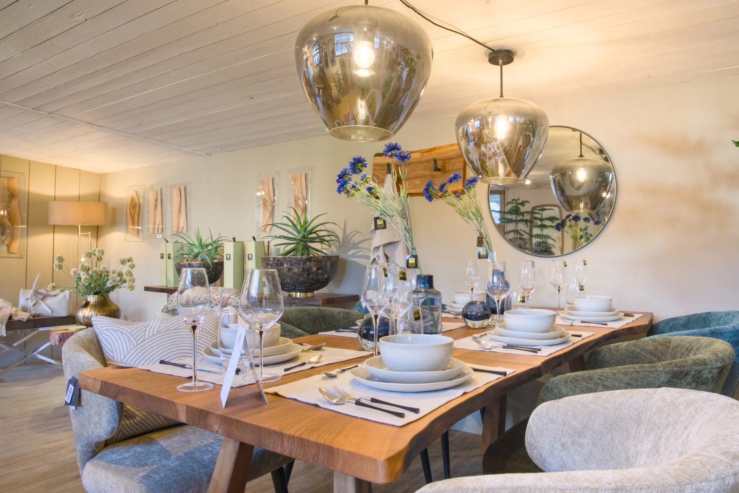 Ein Esszimmer mit einem großen Holztisch, gedeckt mit Speisen, Gläsern und Geschirr, umgeben von gemütlichen Stühlen. An der Wand hängen moderne Lampen, Pflanzen und Spiegel, die den Raum freundlich und einladend wirken lassen.
