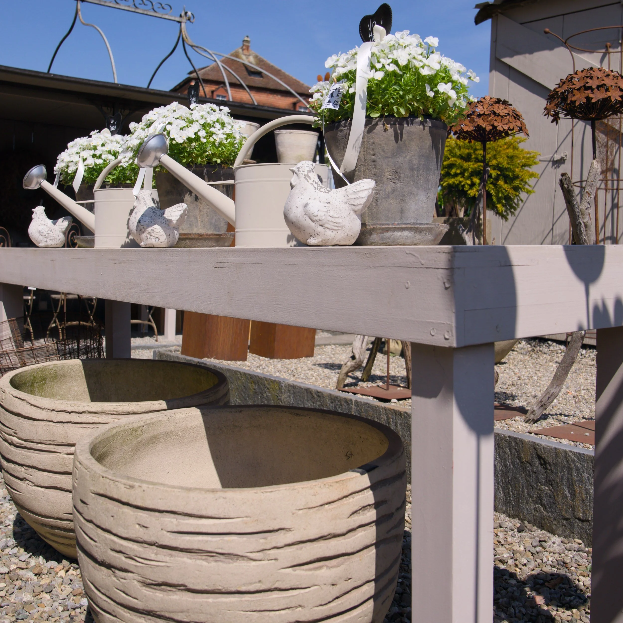 Bunte Blumen in Töpfen, darunter große runde Pflanzgefäße, auf einer Terrasse mit einem weißen Holzgeländer, im Hintergrund ein Gebäude mit einem roten Ziegeldach und blauer Himmel.