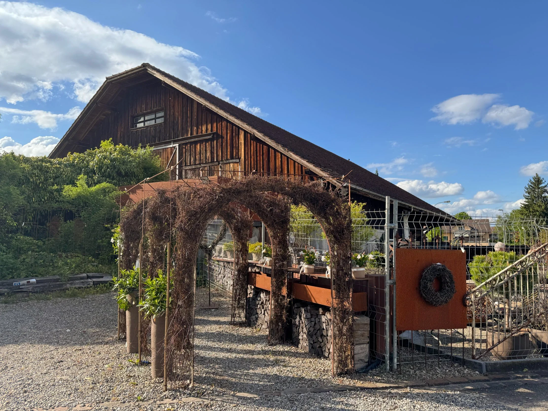 Ein Holzhaus auf einem Hügel, umgeben von Bäumen und Wolken in einem blauen Himmel. Im Vordergrund befinden sich Gartenstrukturen und ein Zaun.