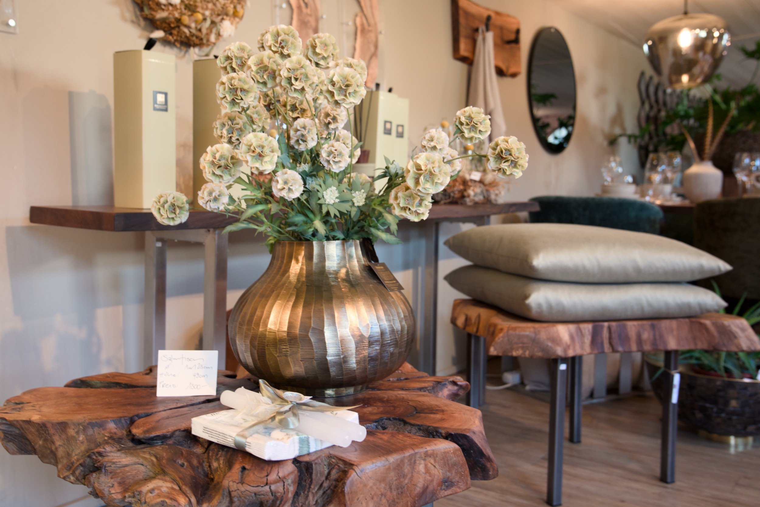 Ein Blumenstrauß mit hellen, großen Blüten in einer goldenen Vase, auf einem rustikalen Holztablett. Im Hintergrund sind weitere Dekorationsgegenstände und Möbel in einem Laden oder Wohnzimmer zu sehen.