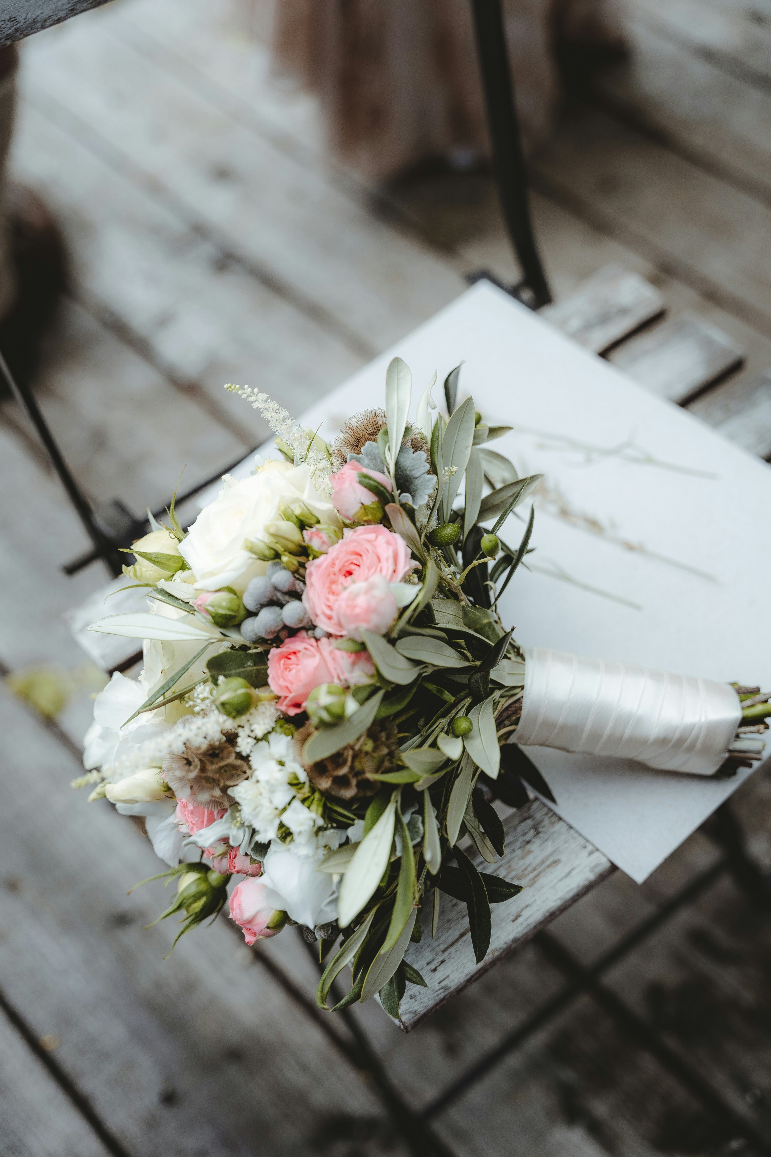Blumenstrauß mit weißen, rosa und grauen Blumen auf einem weißen Tisch, vor einem Holztisch.