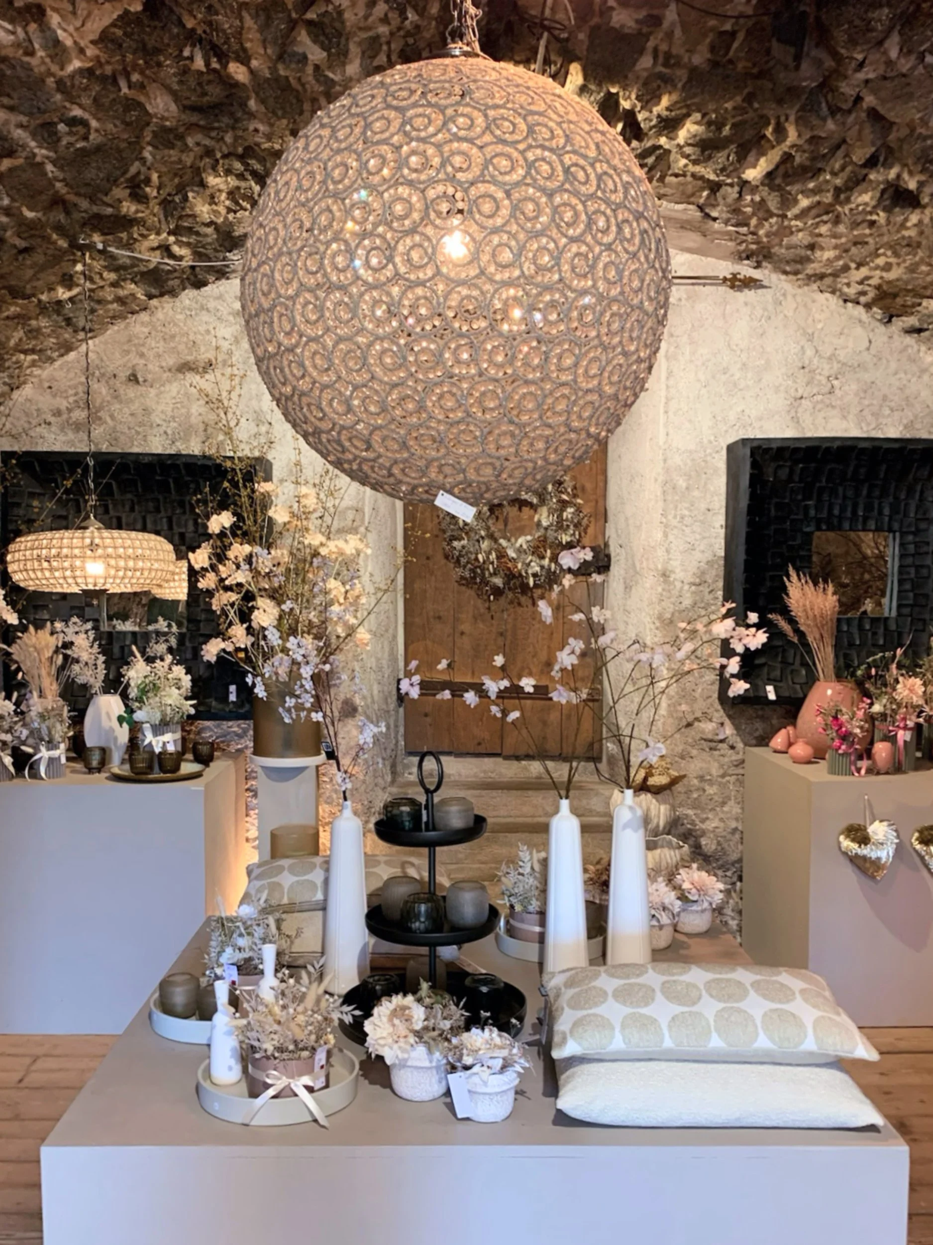 Ein Wohnraum mit rustikalem Steinwand, der mit verschiedenen hellen und neutralen Dekorationsartikeln, Vasen und Kerzen dekoriert ist. Im Mittelpunkt stehen mehrere weiße Vasen, rosa Blumen, Kerzen und Kissen in neutralen Farbtönen. An der Wand hänge
