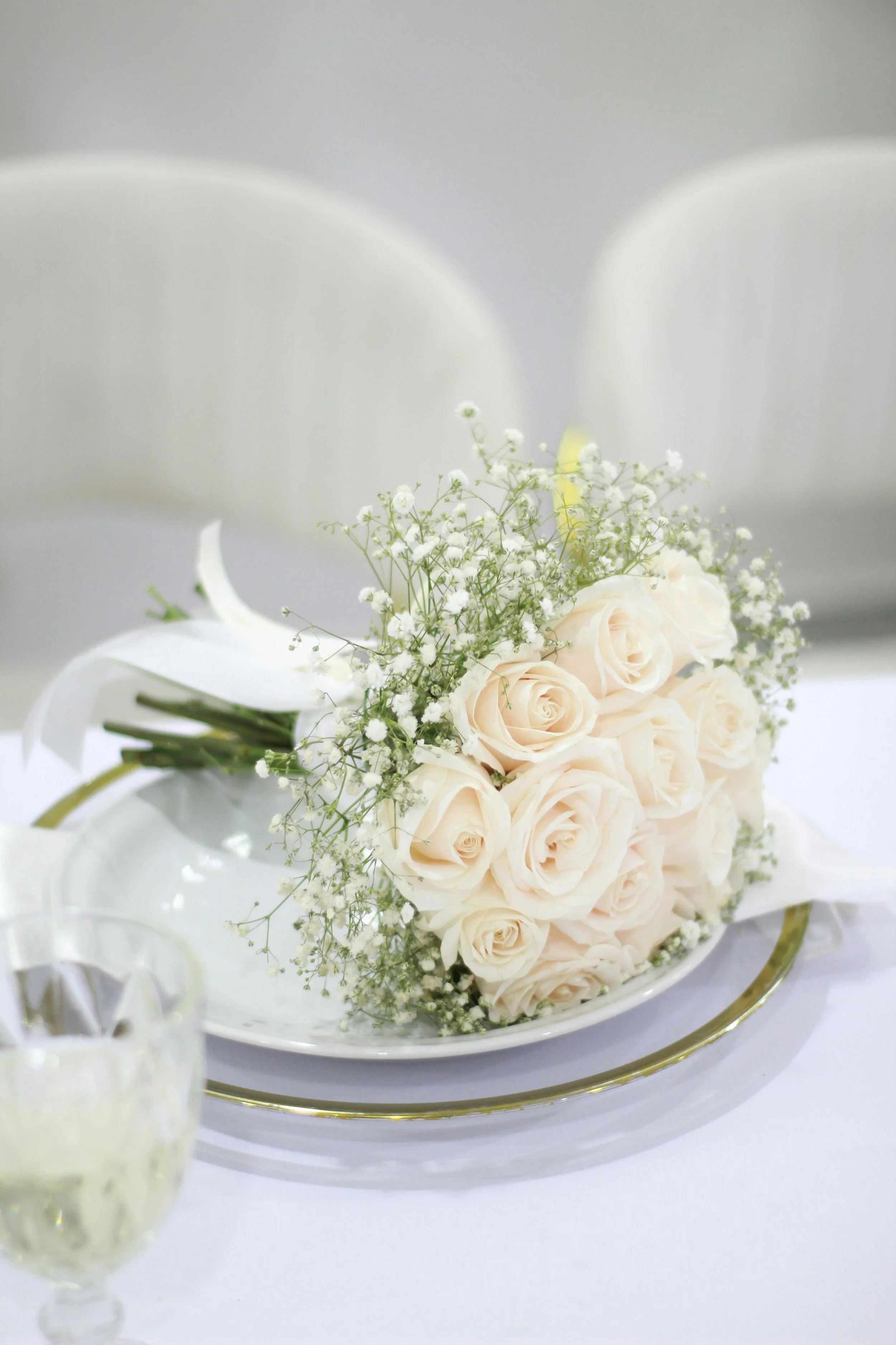 Ein Brautstrauß mit weißen Rosen und kleinen weißen Blumen auf einem weißen Teller, im Hintergrund zwei weiße Stühle.