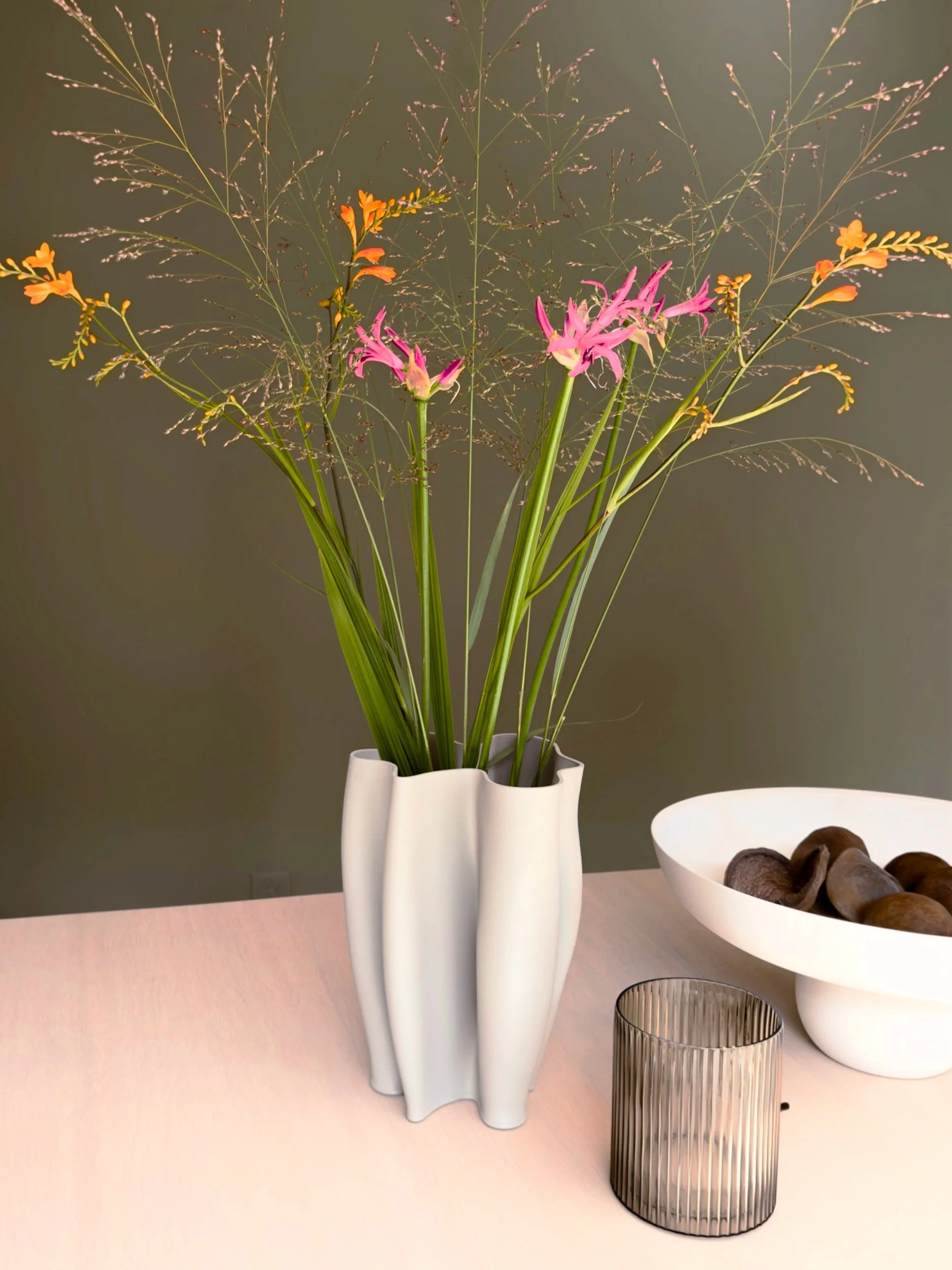 Eine weiße Vase mit dünnen grünen Gräsern und pink-orangefarbenen Blumen steht auf einem hellen Tisch, daneben eine leere, silberne Trinkglas und eine weiße Schale mit dunklen, runden Gegenständen.