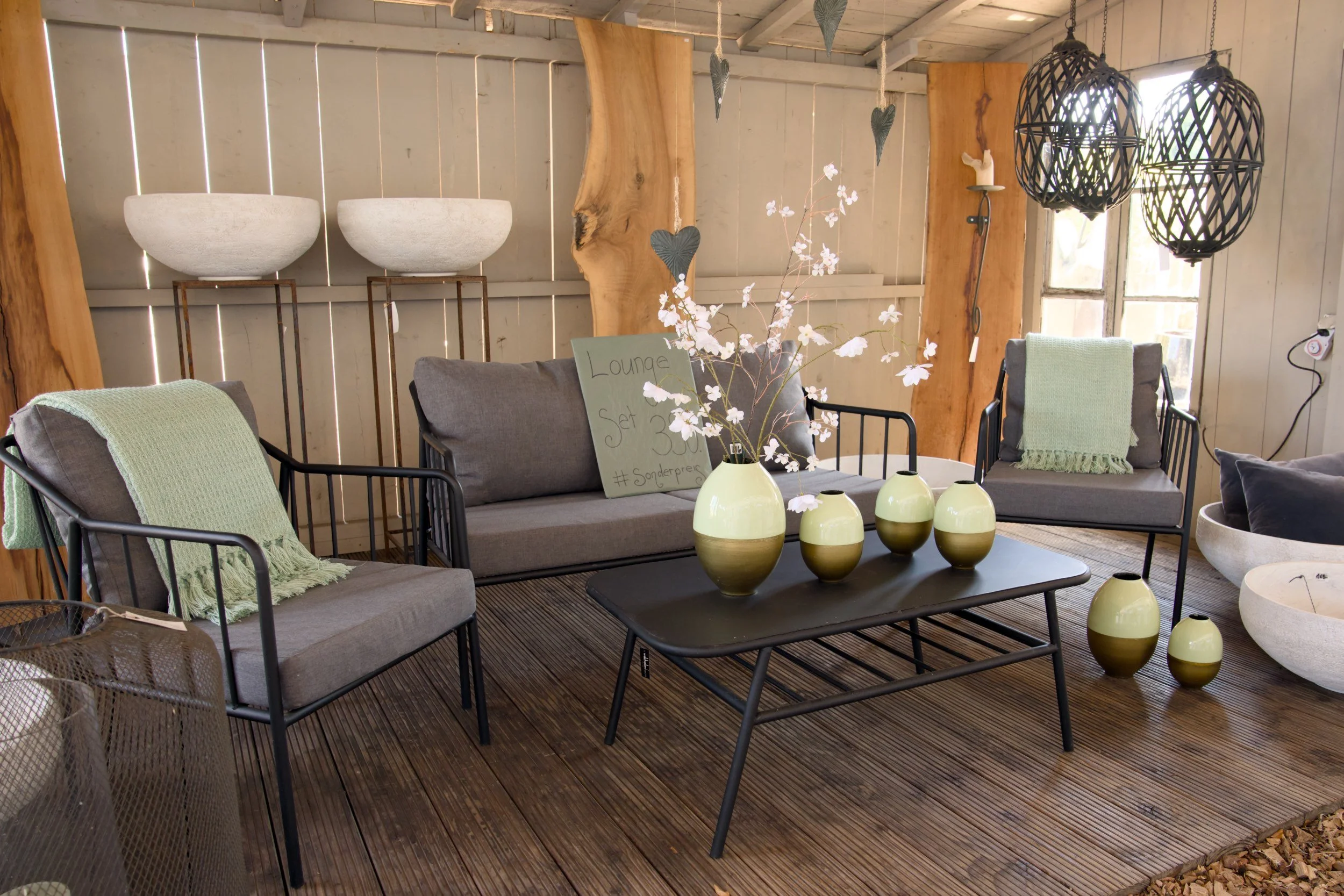 Gemütliches Wohnzimmer mit grauen Sesseln, einer Kaffeetafel mit weißen Vasen und einer Blume, sowie dekorativen Lampen und Holzdekoration an den Wänden.