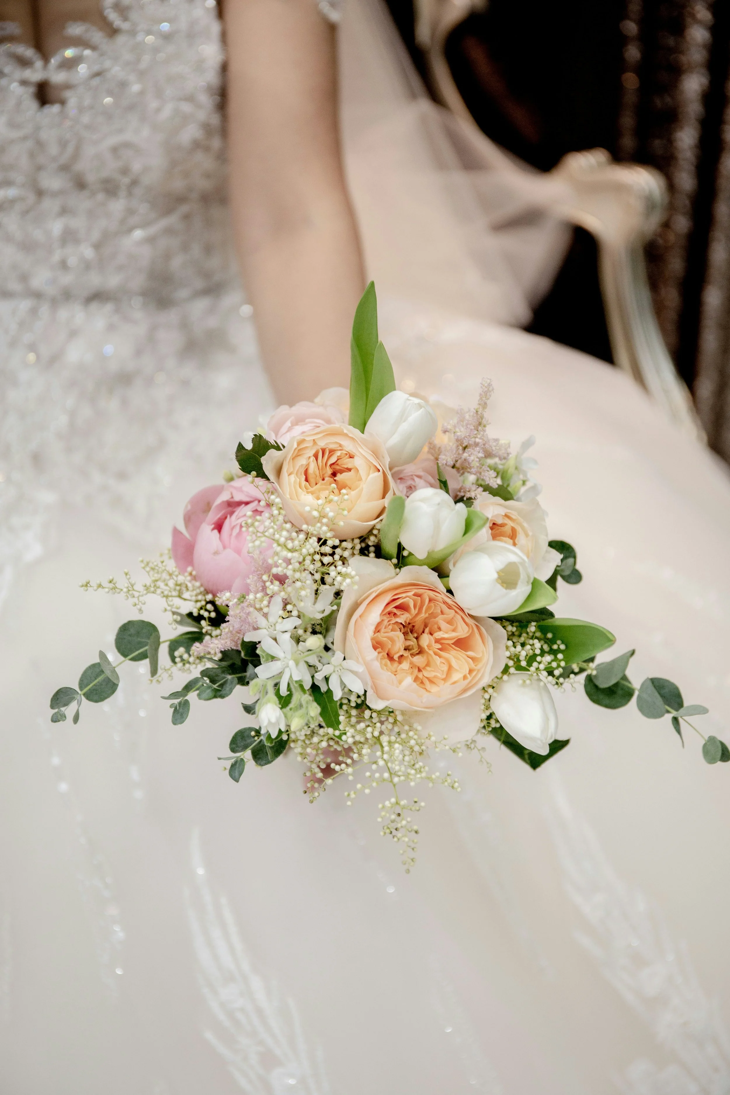 Ein Brautstrauß mit weißen Tulpen, peachfarbenen Rosen, kleinen weißen Blumen und grünem Eukalyptus auf einem weißen Untergrund.