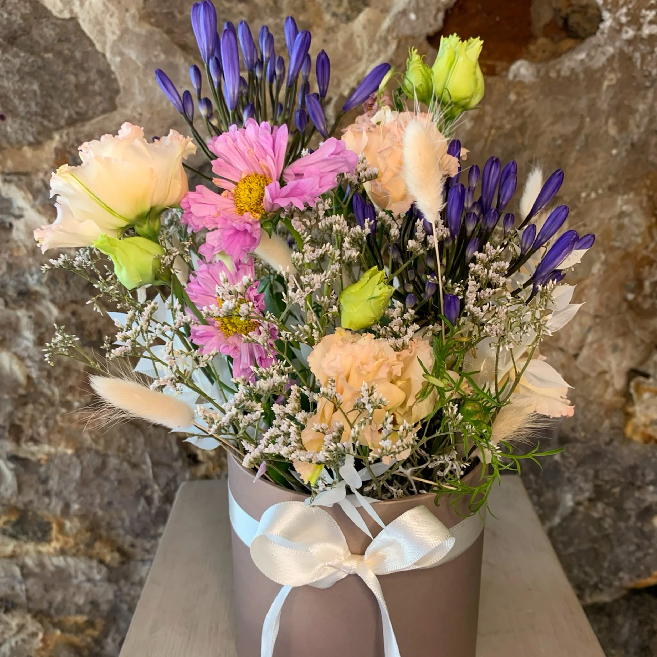 Blumenstrauß mit lilafarbenen, rosa, cremefarbenen und weißen Blumen sowie Federn, mit brauner Steinwand im Hintergrund.