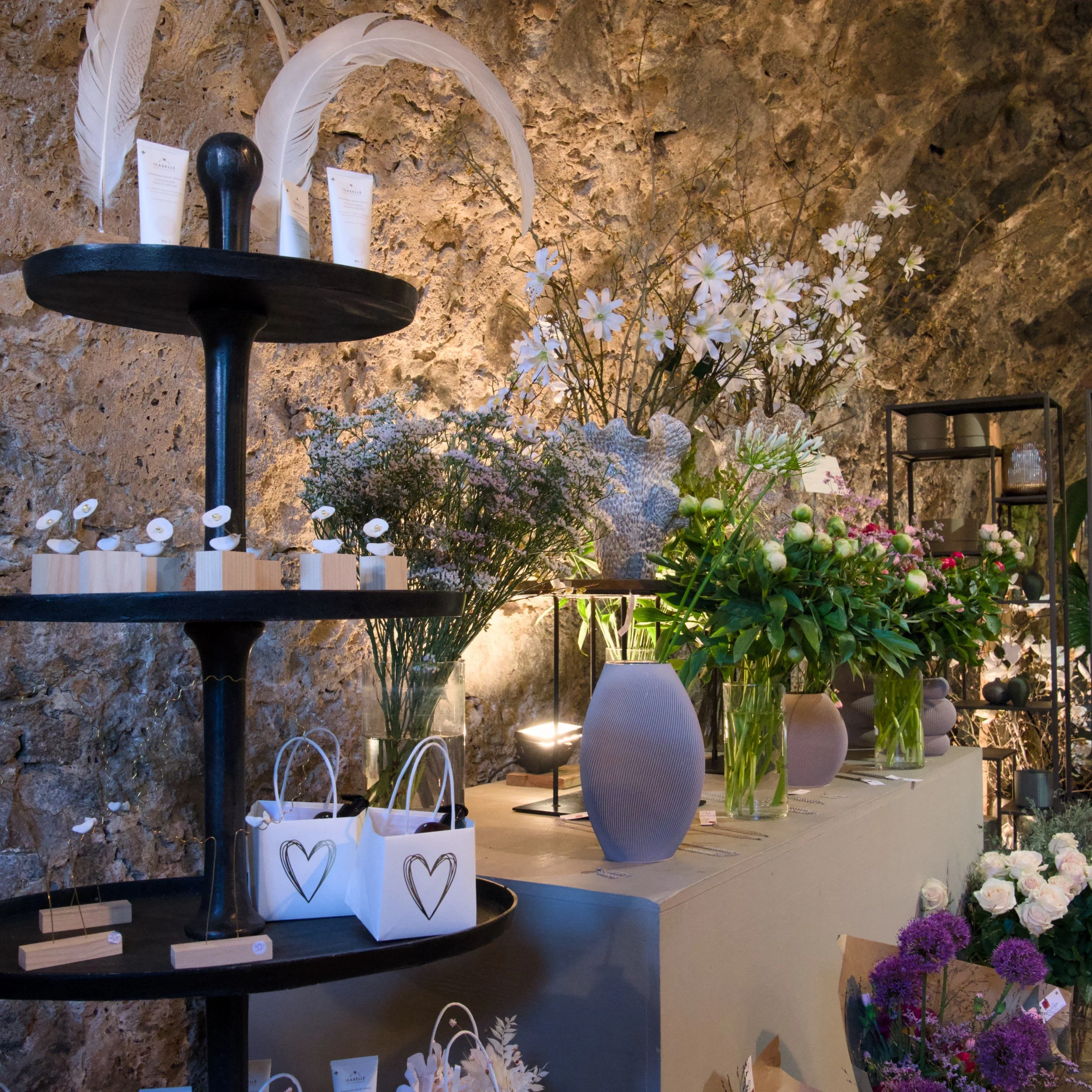 Blumen aus verschiedenen Sorten in Vasen und Körben, eine schwarze dekorative Figur, eine schwarze Tafel auf einem Tisch, ein Regal mit Kerzen und Gegenständen, eine Steinumwand im Hintergrund.