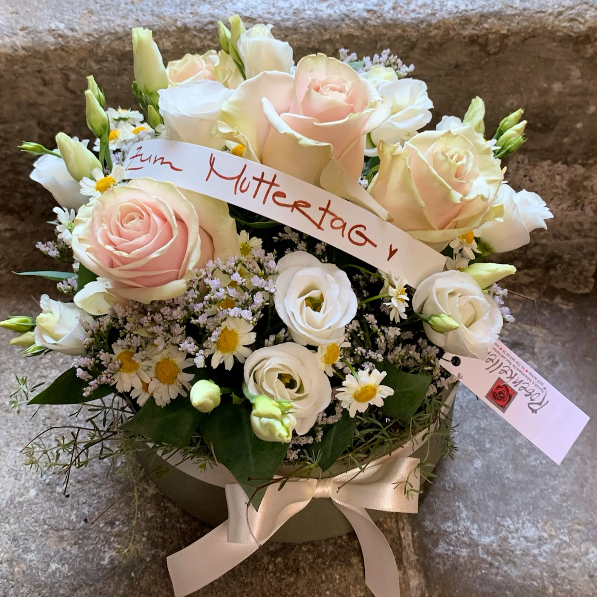 Blumenstrauß mit weißen und pastellfarbenen Rosen, kleinen weißen Blumen und einem Geschenkband mit einem Schild, das Liebe ausdrückt, vor einem grauen Steinboden.