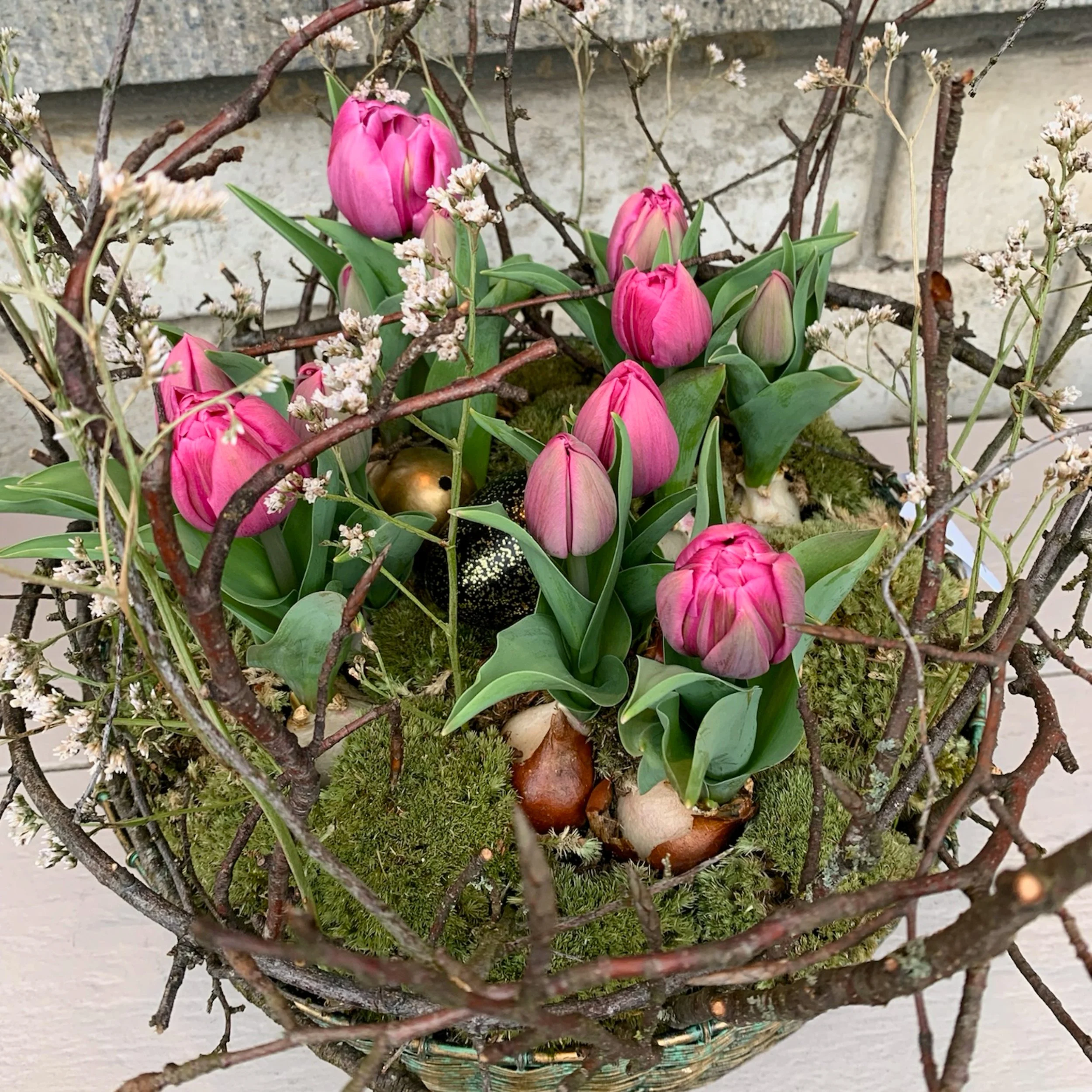 Ein Korb mit pinkfarbenen Tulpen, grünen Blättern, kleinen weißen Blumen, Moos und dekorativen Eiern, umgeben von Zweigen.