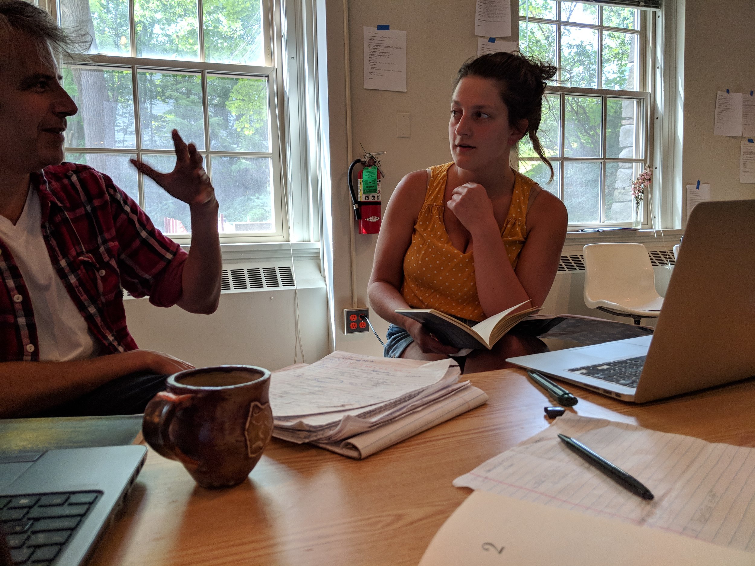 Two people having a conversation at a table in a room with large windows. One person is gesturing while speaking, the other is listening and holding a book. There are papers, a laptop, and pens on the table.