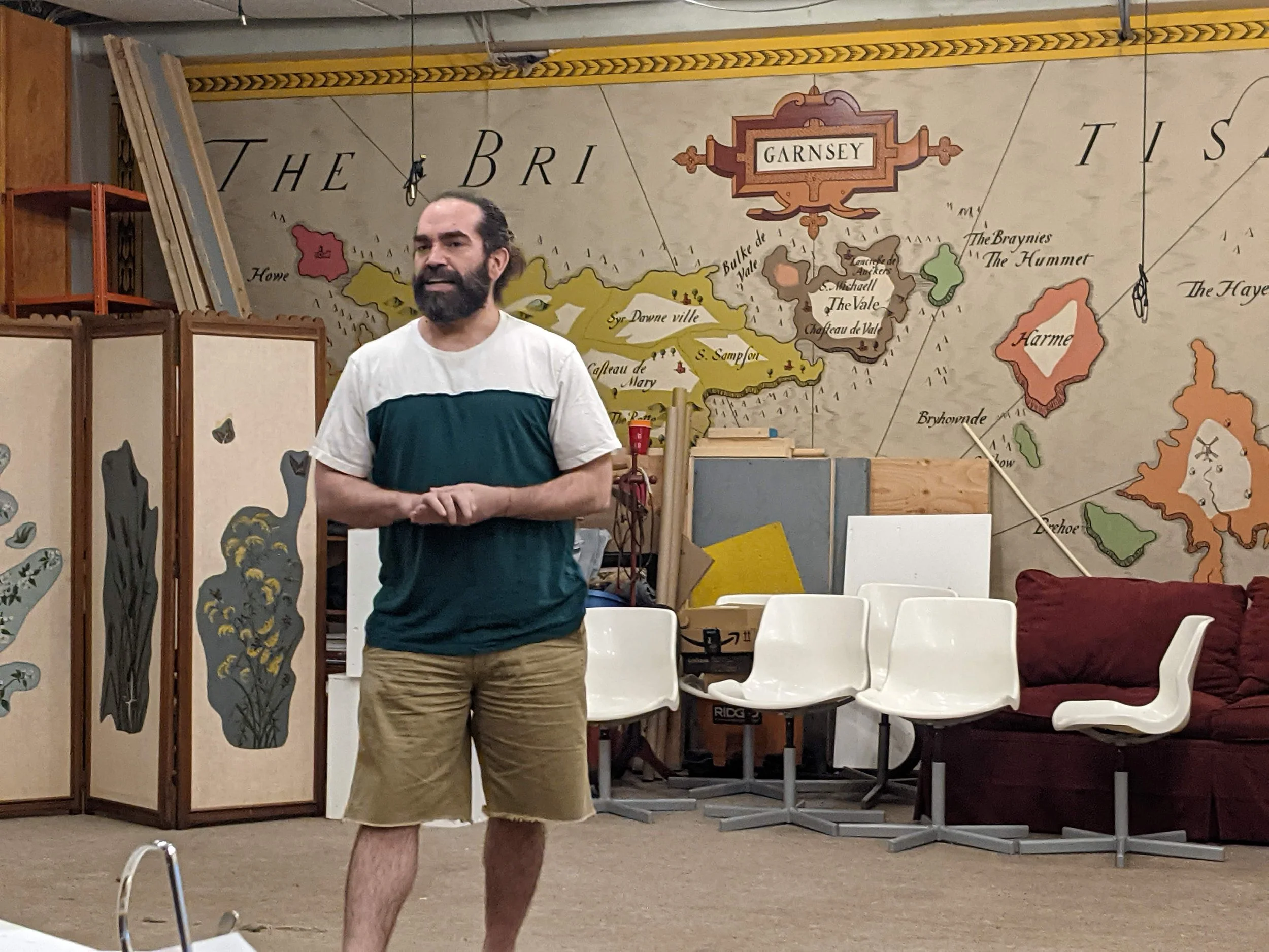 A man with a beard and long hair tied back, wearing a white and dark green t-shirt and tan shorts, standing in a room with a large map on the wall behind him, white chairs, a red couch, and a folding room divider with a floral design.