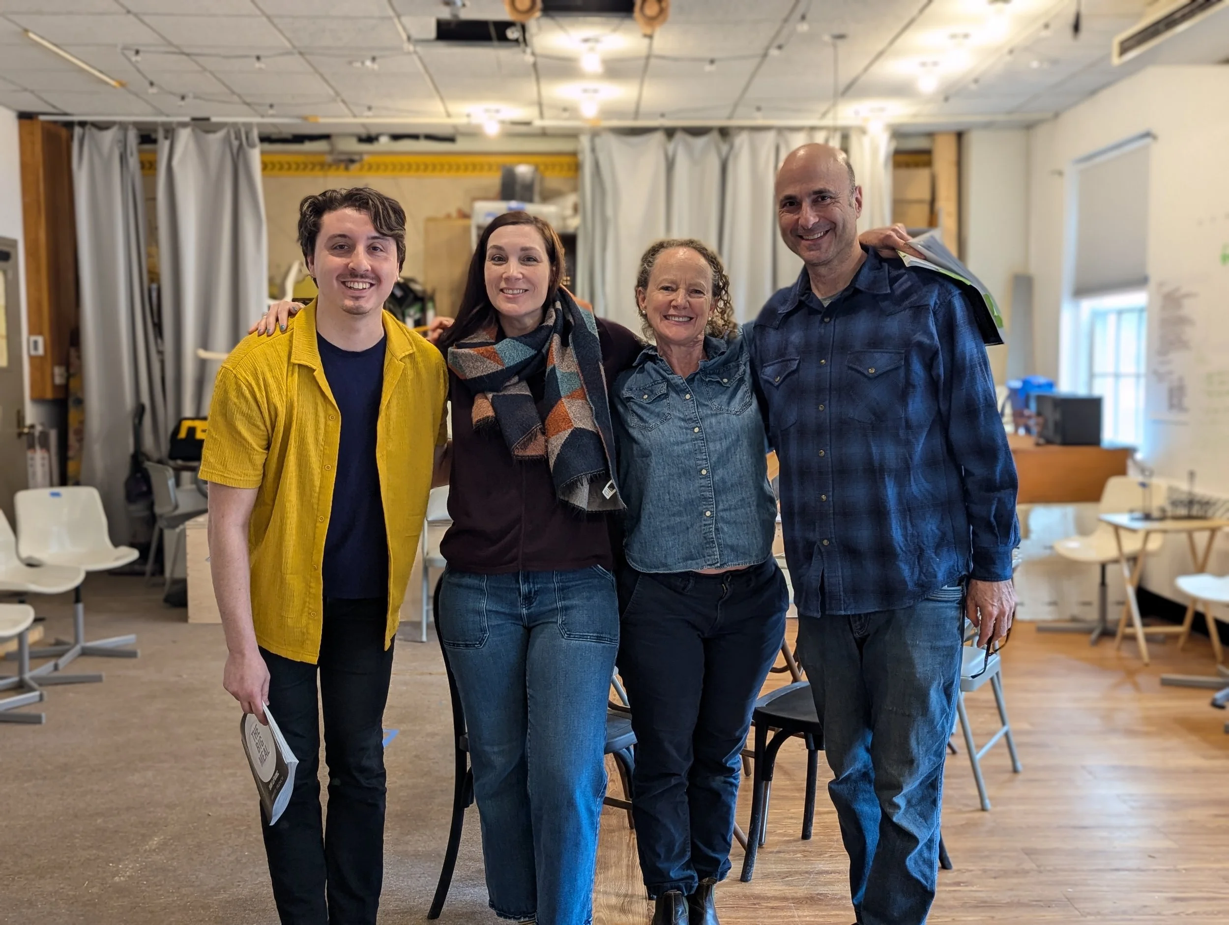 Four people standing indoors, smiling and posing for the camera, in a room with chairs and a whiteboard in the background.
